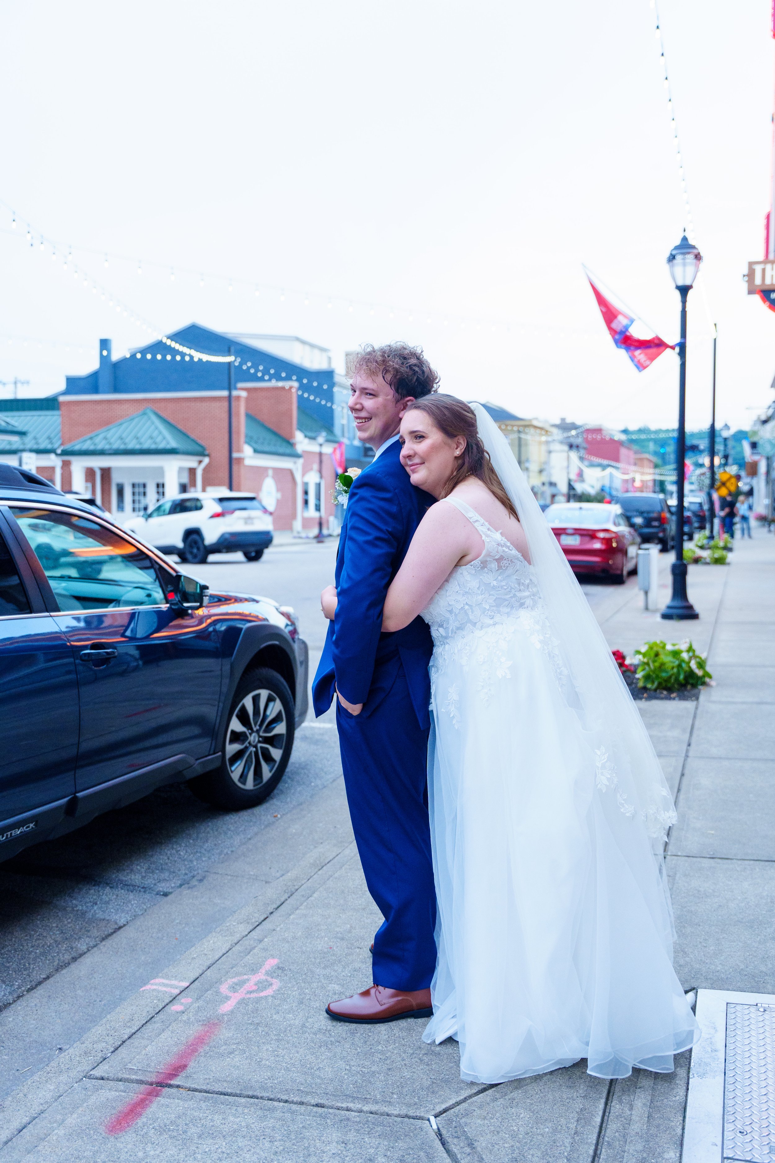 A happy bride and groom sharing a moment on a city street during their wedding, with colorful buildings, parked cars, and decorative string lights in the background.