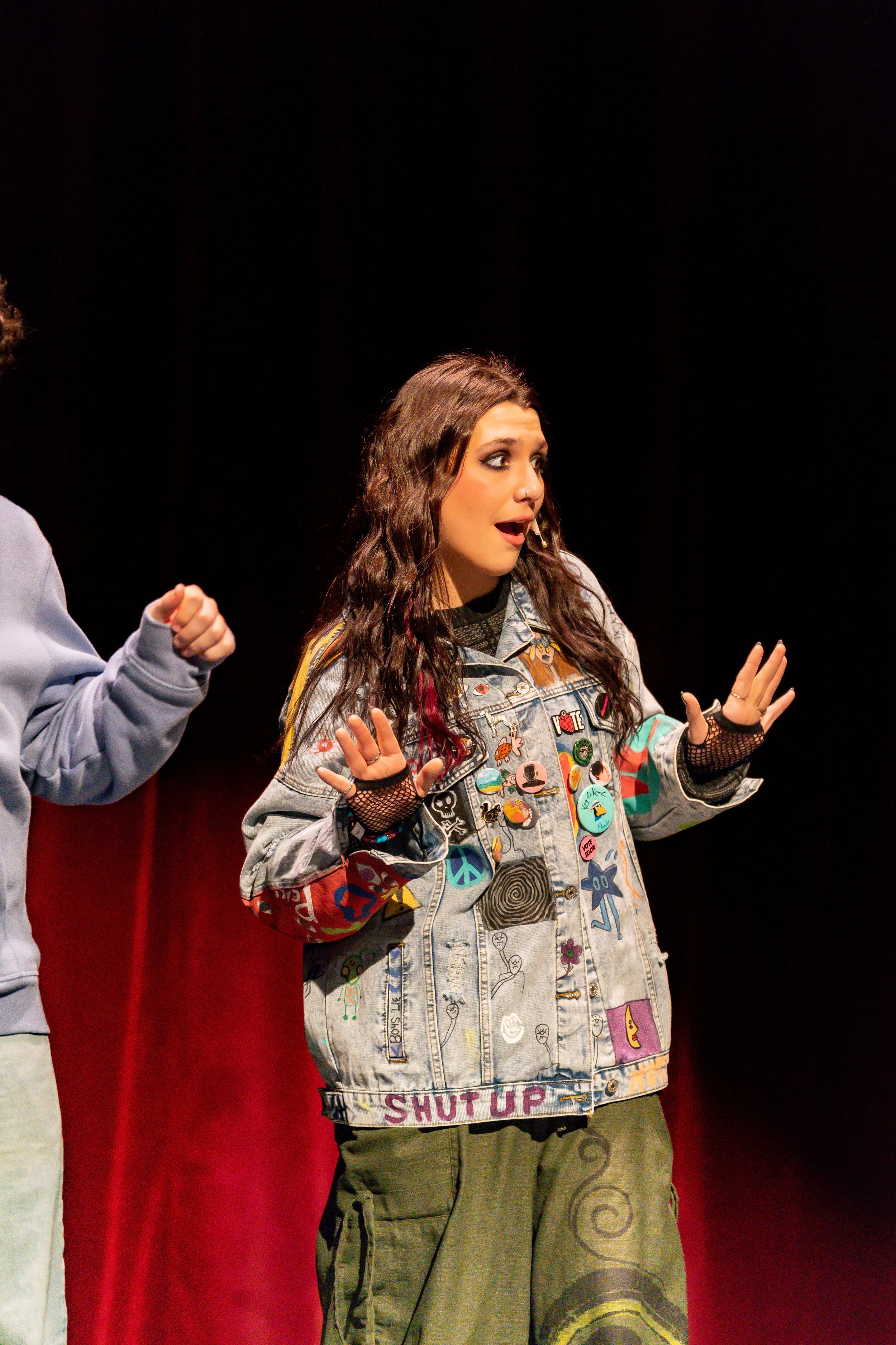 A woman with long dark hair and a surprised expression, wearing a denim jacket with colorful patches and pins, standing in front of a red curtain.