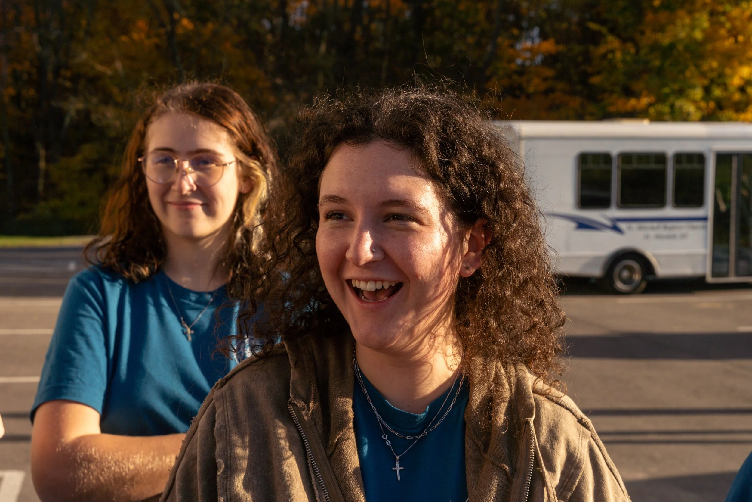 Two young women standing outdoors, one with curly hair and a cross necklace, and another with glasses and wavy hair, smiling with a bus and autumn trees in the background.