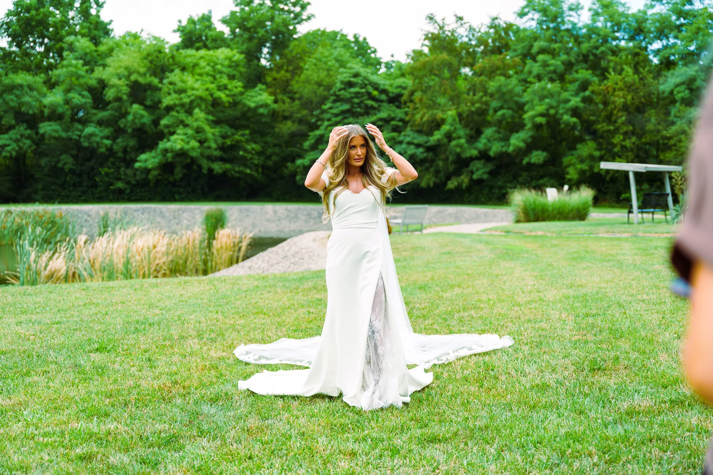 A woman in a white wedding dress standing on a grassy area in a park with green trees in the background, holding her head with both hands.