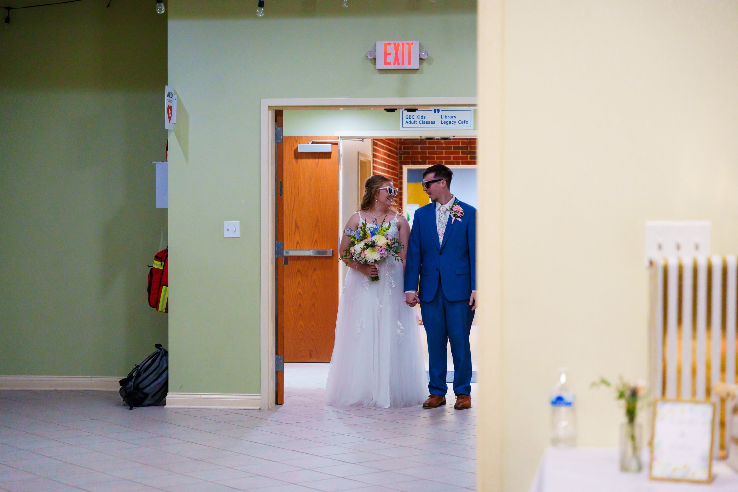 A bride and groom holding hands walking into a room. The bride is wearing a white wedding dress with a floral bouquet, and the groom is dressed in a blue suit with sunglasses.