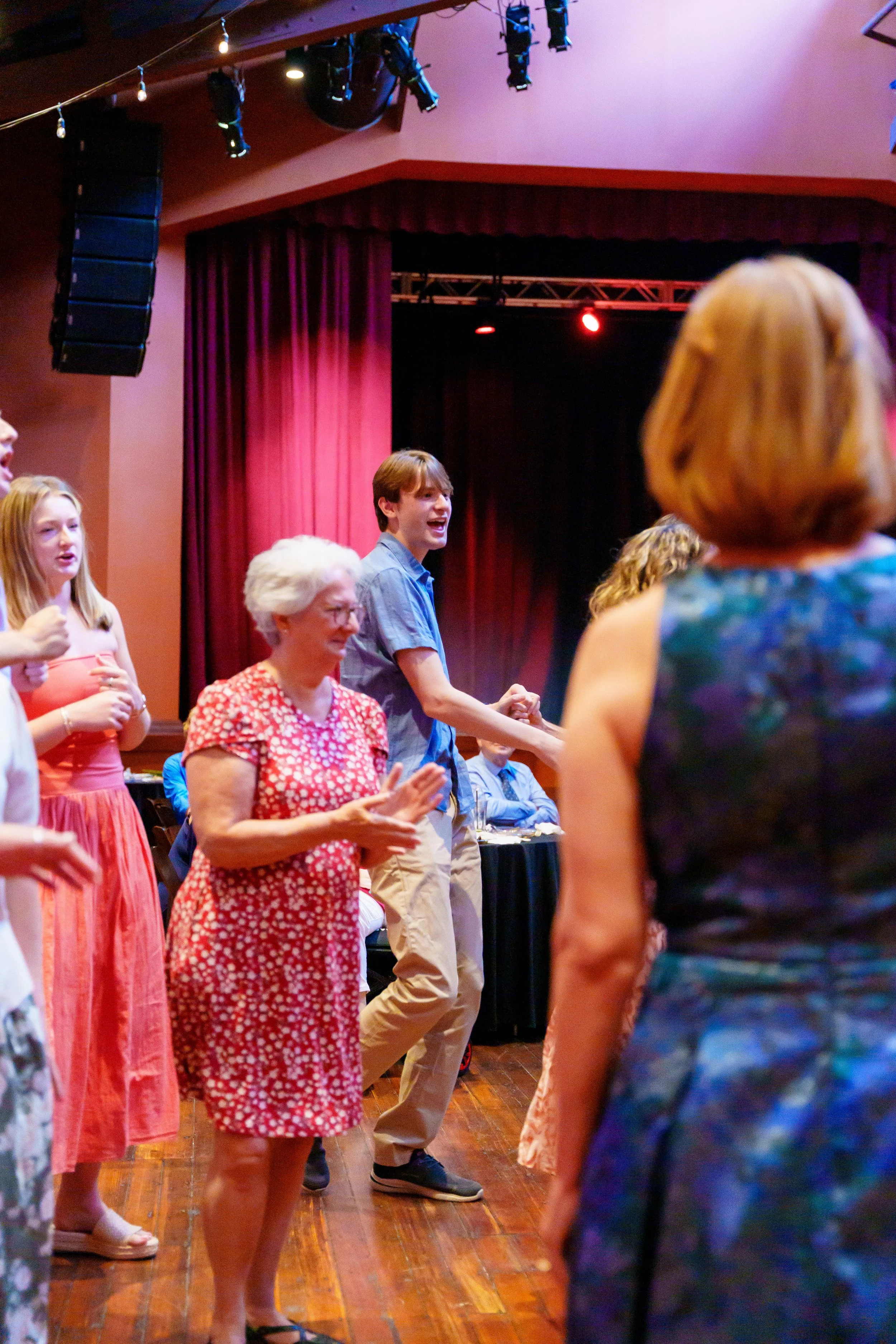 People standing in a line on a wooden stage or floor, holding hands, with a red curtain and black stage equipment in the background, participating in a group activity or dance.