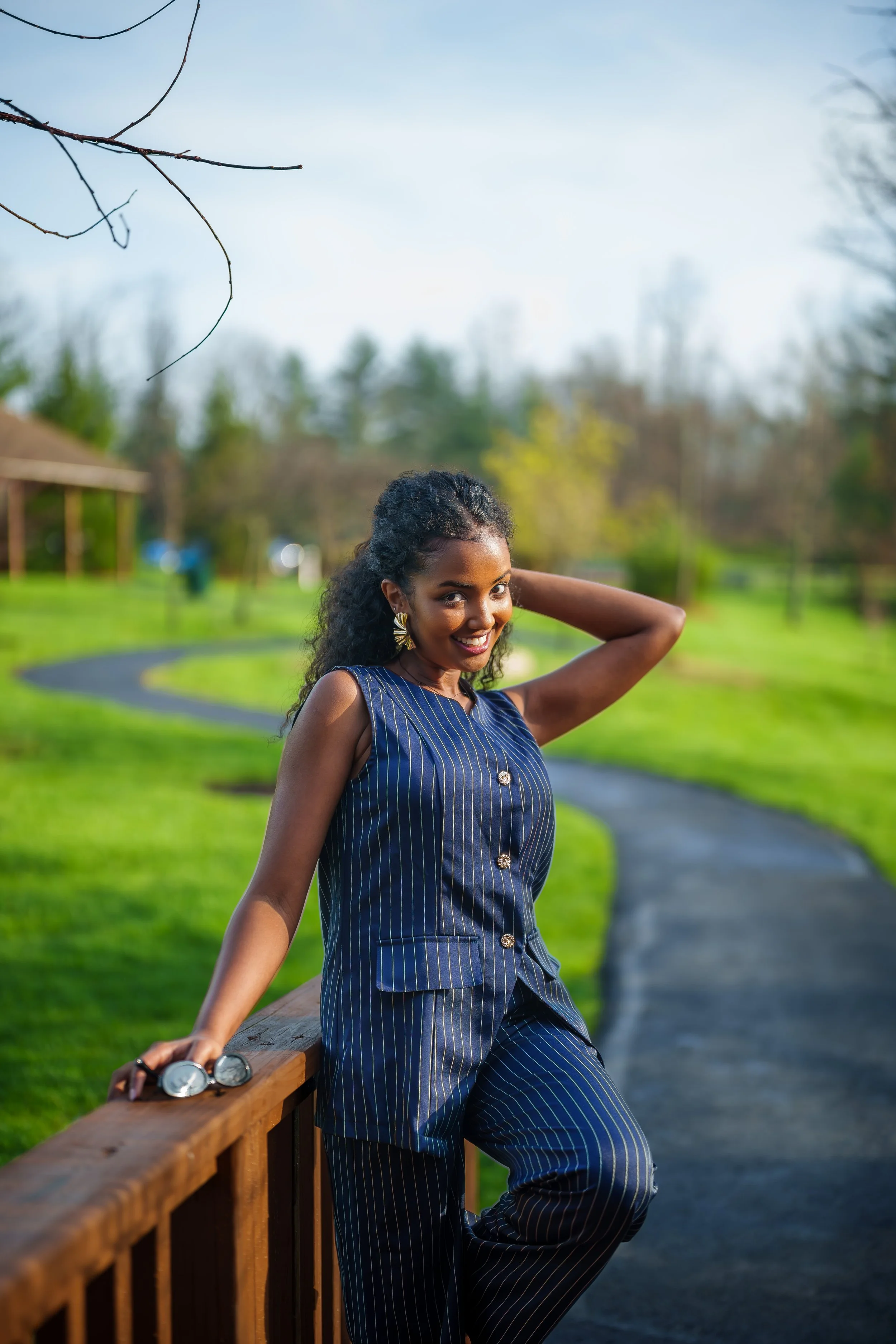 A woman in a blue pinstriped sleeveless outfit with gold buttons and earrings, smiling and leaning on a wooden railing in a park with green grass, trees, and a winding pathway.