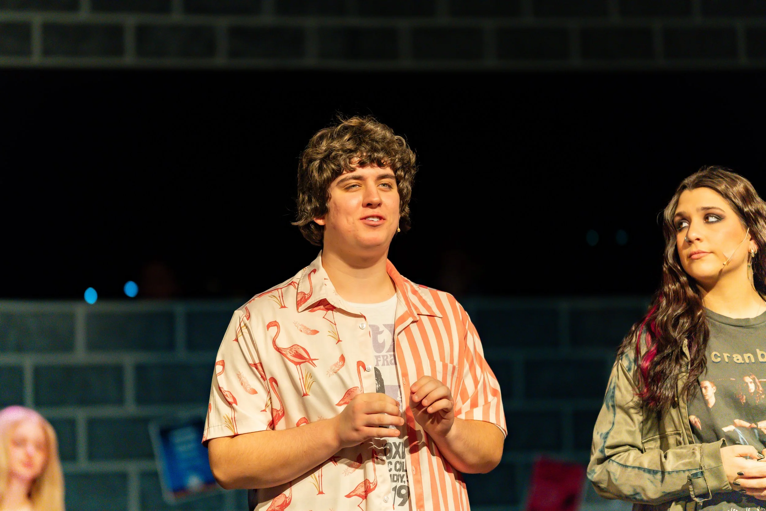 A young man with curly hair standing in front of a black background, wearing a shirt with flamingo and palm tree patterns, and another woman with long dark hair and a face mask, standing beside him, in an outdoor setting at night.