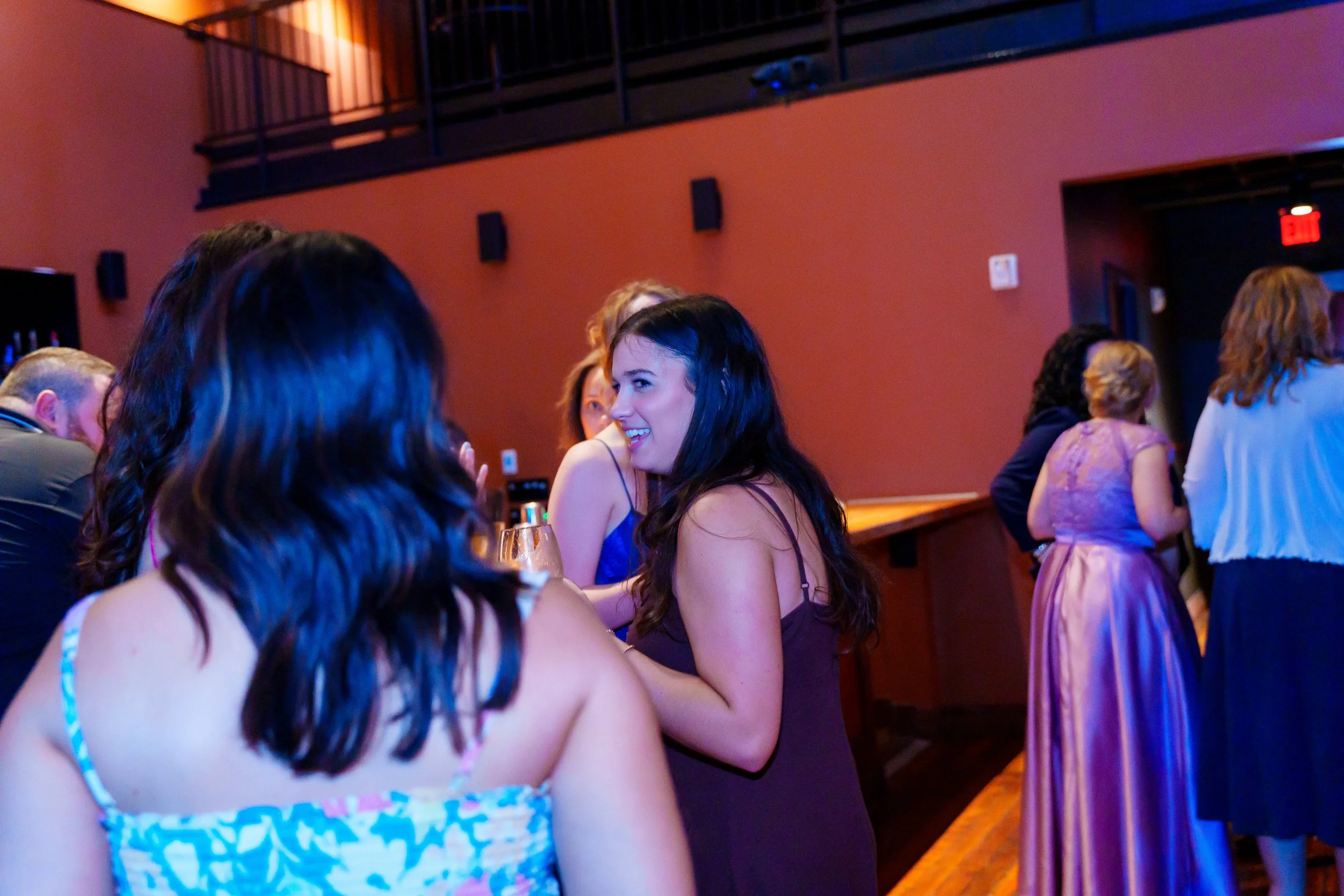 Group of people socializing in a dimly lit bar or lounge, with women talking and smiling, some wearing dresses, and a bar area visible in the background.