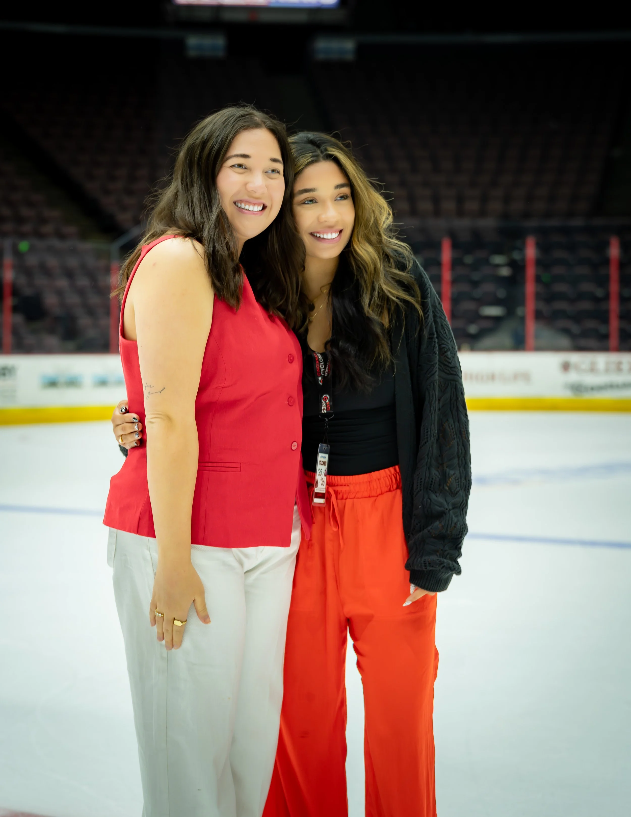 Two women smiling on an ice rink, one in a red top and white pants, the other in a black top and orange pants, with empty arena seats behind them.