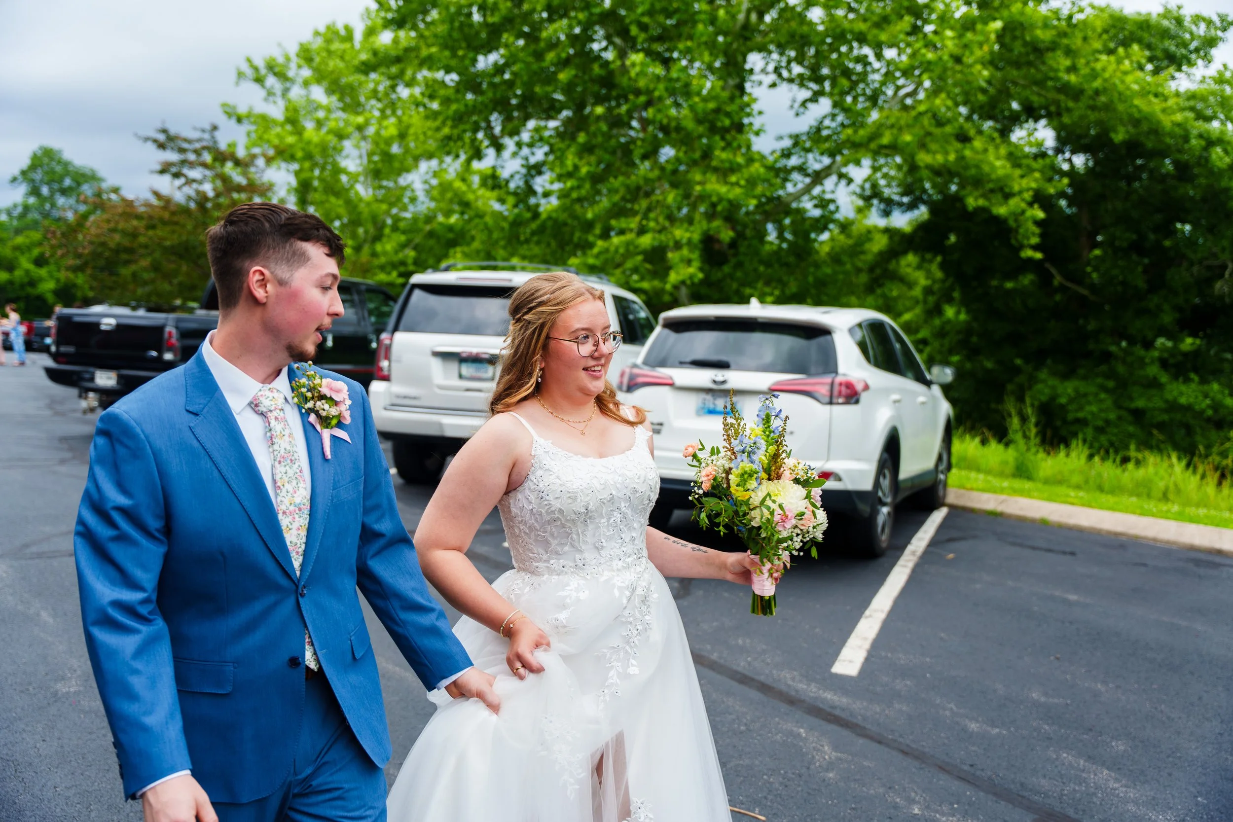 A bride and groom walking in a parking lot with trees in the background. The bride is holding a bouquet of flowers and wearing a white wedding dress, while the groom is in a blue suit with a floral tie and boutonniere.