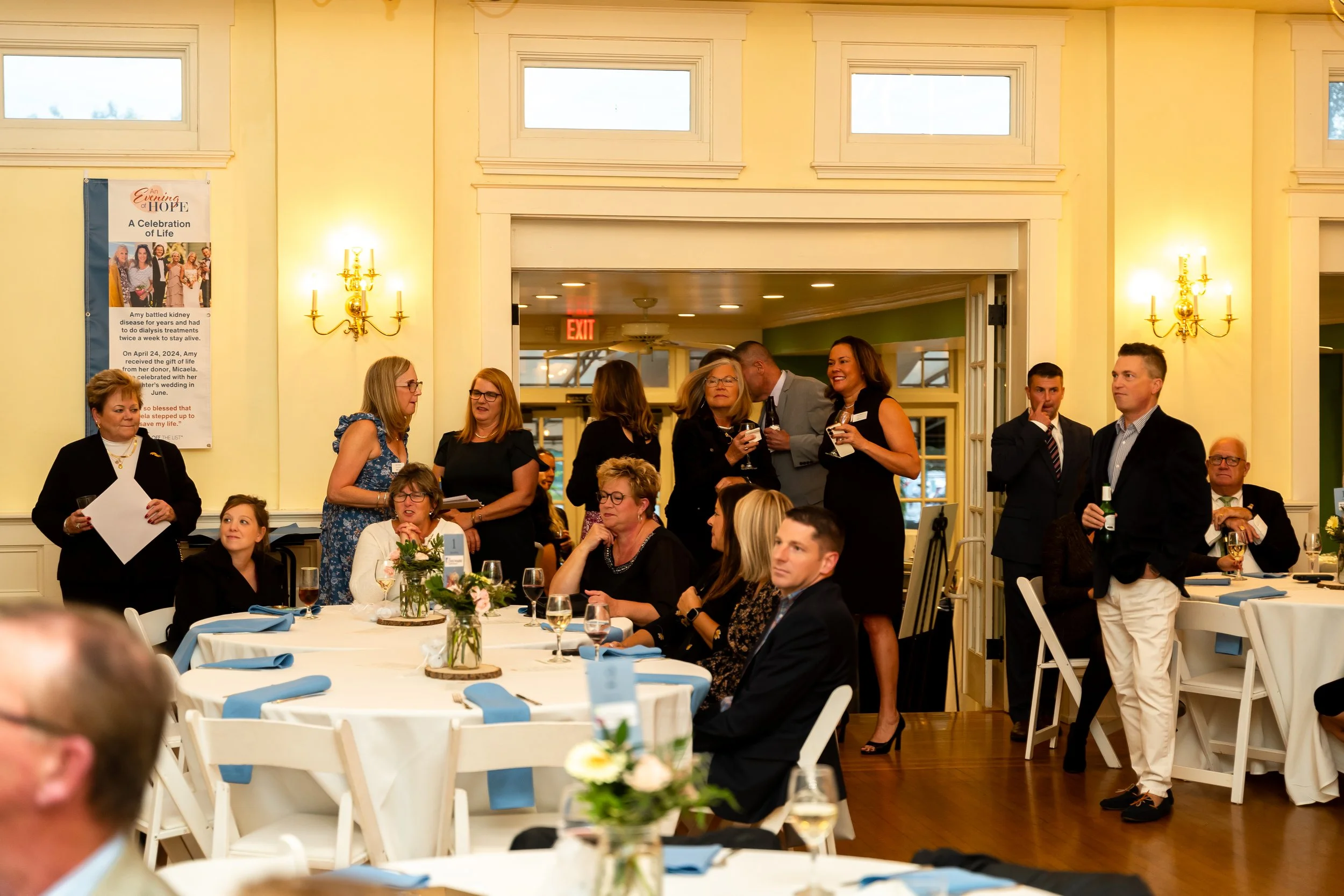 People socializing and talking at a formal event or banquet in a decorated hall with yellow walls and high windows.
