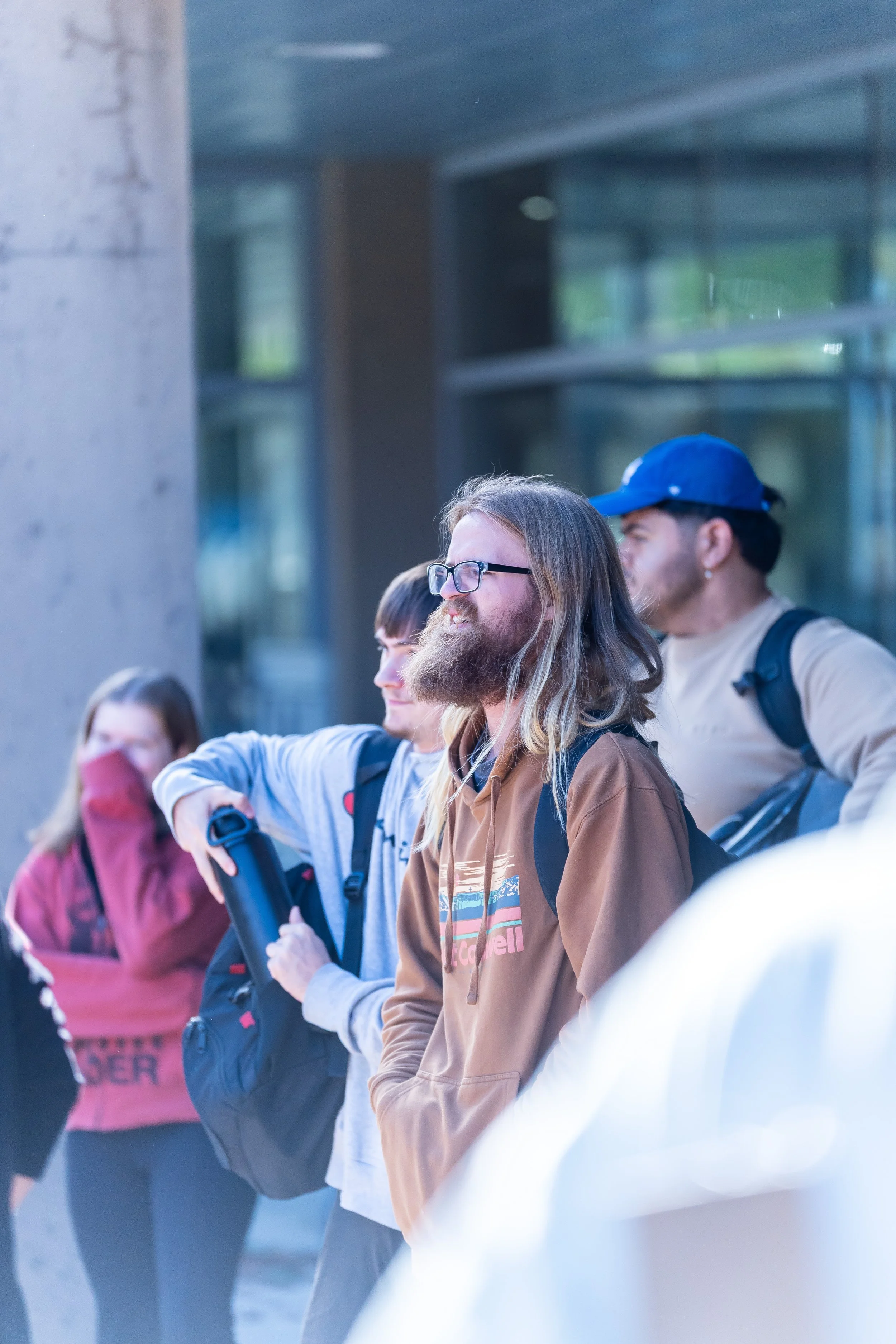 A group of people standing outdoors, some wearing backpacks, in front of a modern building with large glass windows.