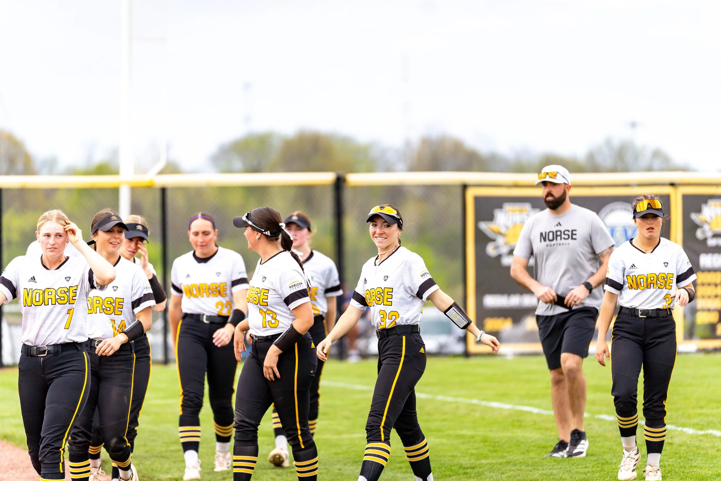 A group of female softball players from the Norse team walking together on a field. They are wearing white jerseys with yellow and black accents, and black pants with yellow stripes. Some players are wearing visors or hats, and one has sunglasses. Th