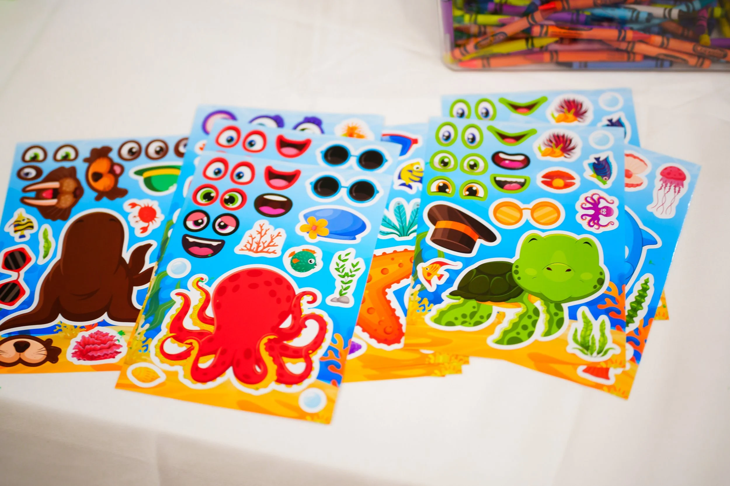 Colorful ocean-themed sticker sheets with sea creatures, smiling faces, coral, and seaweed, laid out on a white surface with a plastic container of crayons in the background.