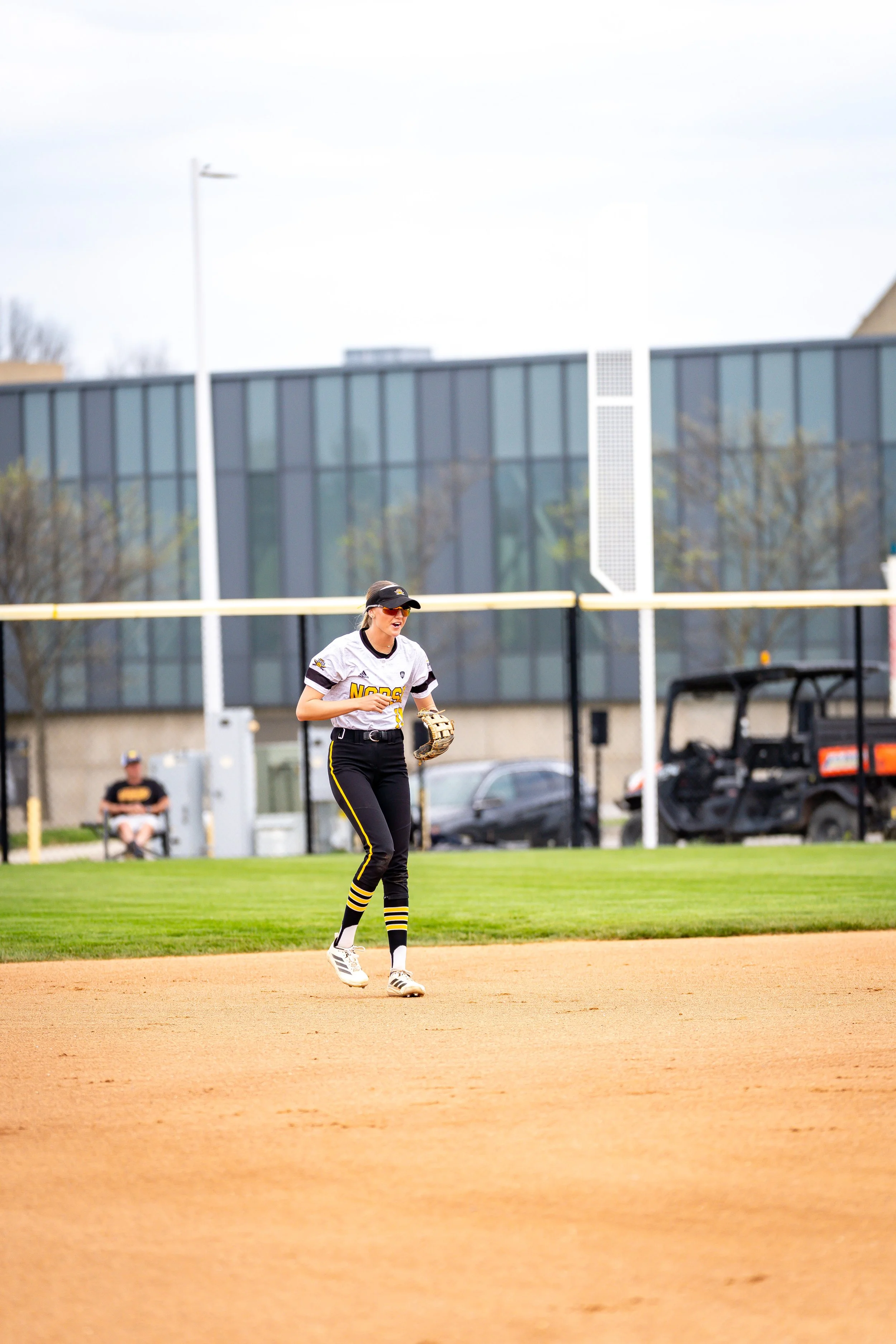 A female softball player wearing a white and black uniform and a black cap is standing on a baseball field near the baselines, with a grassy outfield, a building with large glass windows, and a fence in the background.