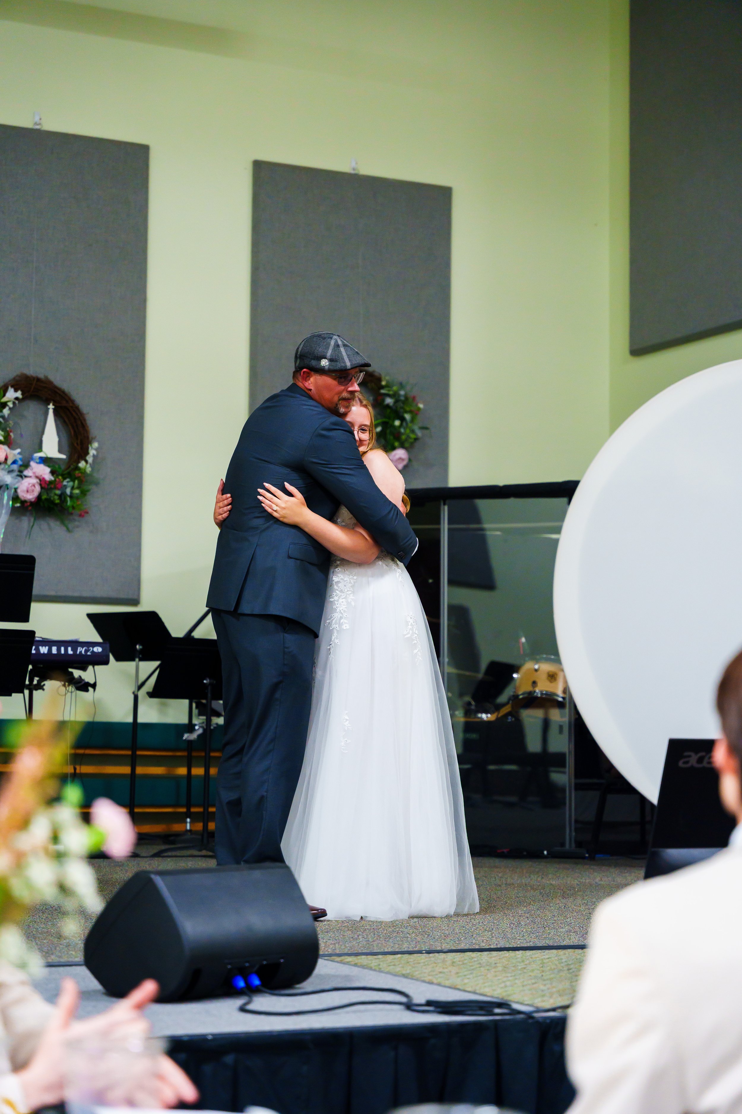 A man in a dark suit and flat cap hugging a woman in a white dress on stage at a wedding or formal event. Decorations and musical instruments are visible in the background.
