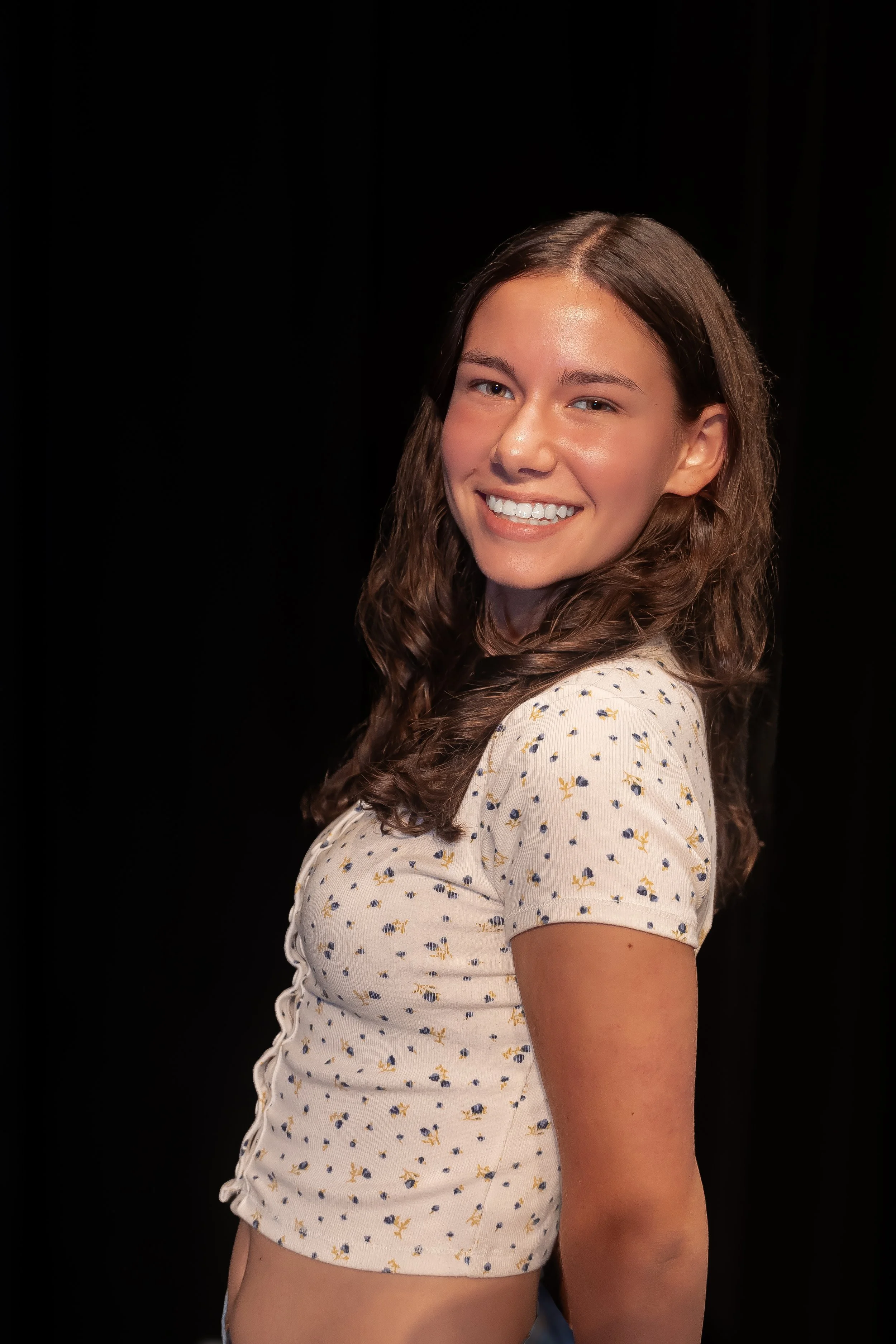 A young woman with long wavy brown hair smiling at the camera, wearing a cream-colored shirt with a yellow and blue floral pattern, against a black background.