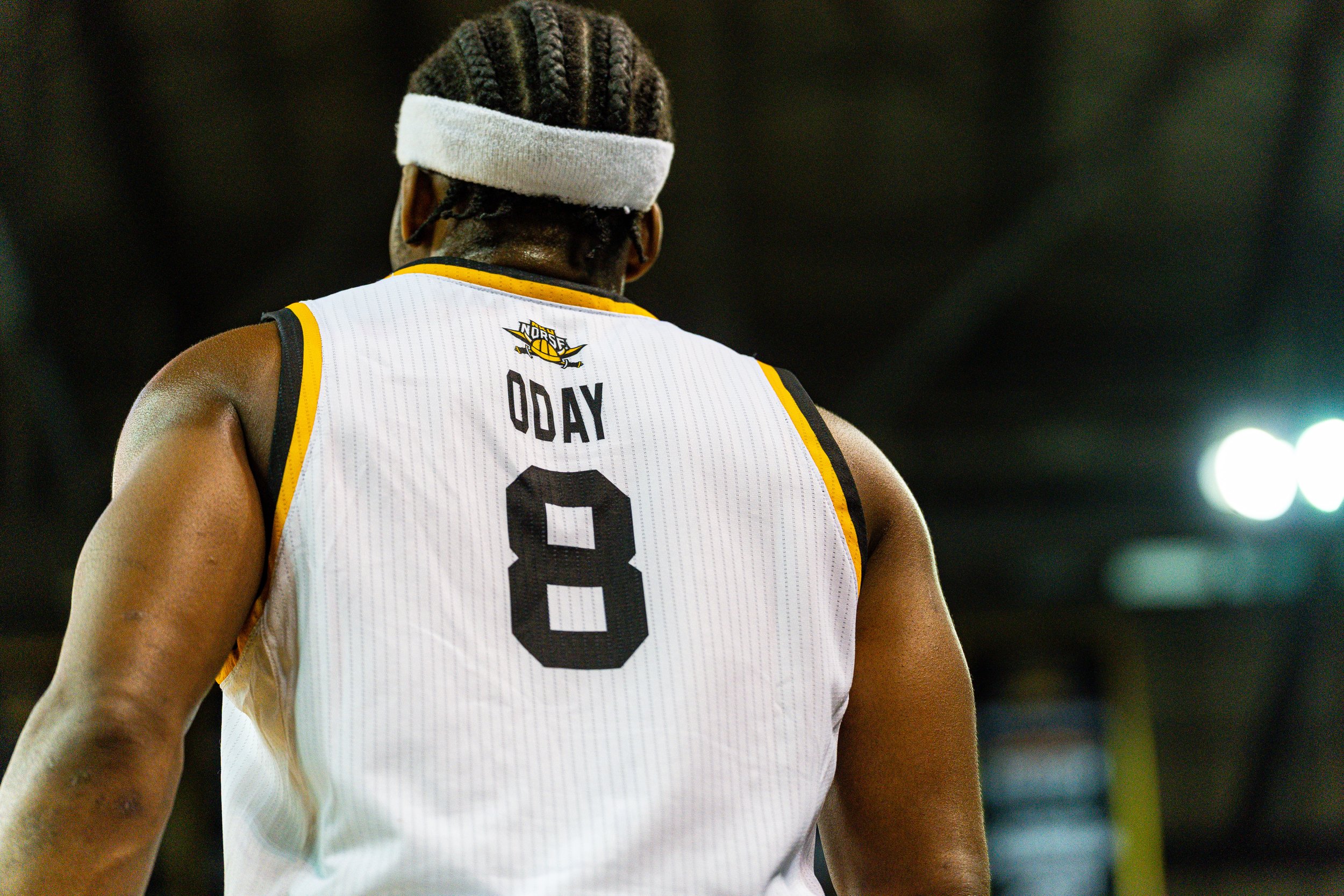 Back of a basketball player wearing a white jersey with yellow and black accents, the number 8 on it, and the name 'Oday'; he has a headband and braided hair, and is facing away from the camera with a dark, blurry background.