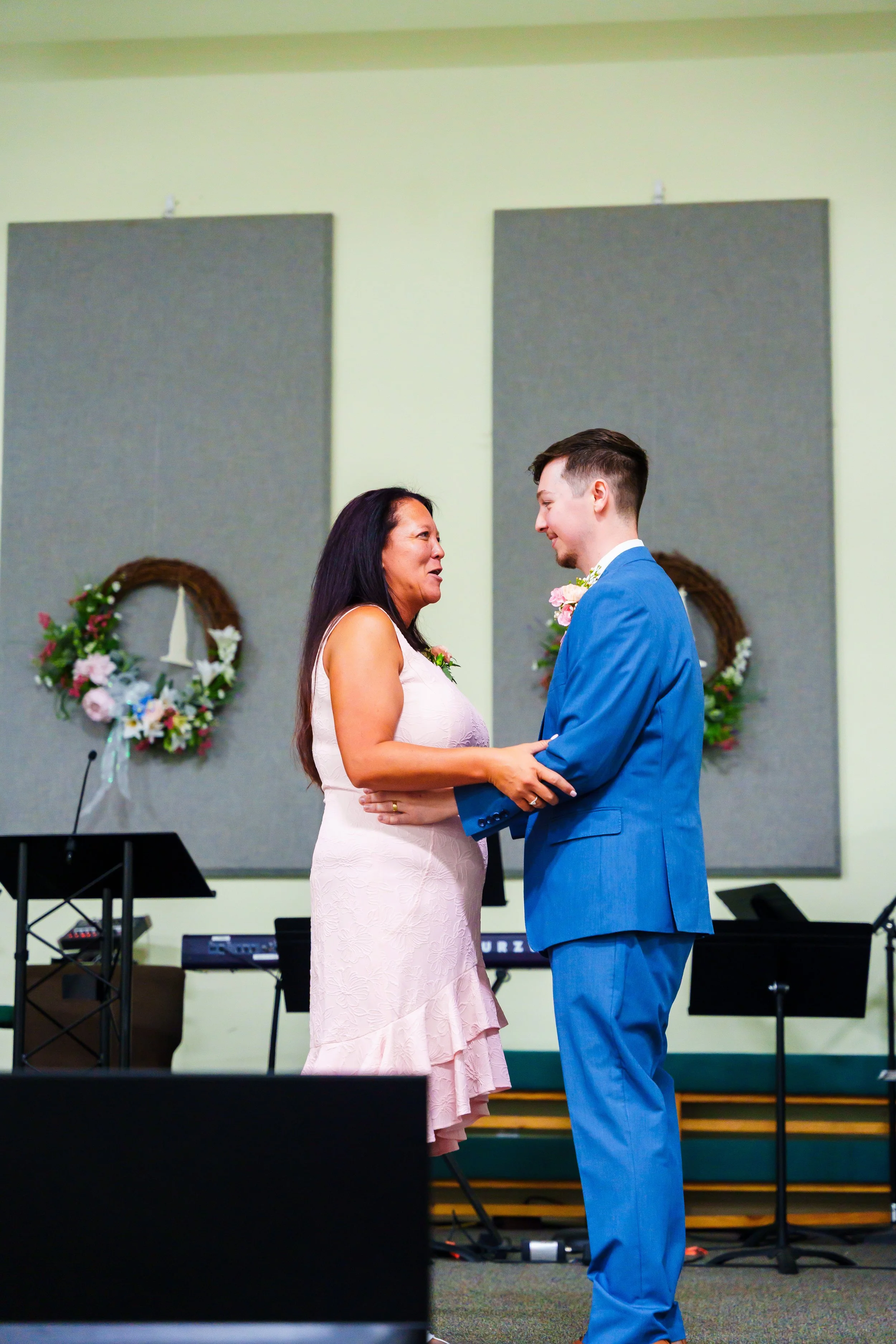 A woman and a man are standing face to face in a church, holding hands, during a wedding ceremony. The woman is wearing a light pink dress and the man a bright blue suit, both smiling.