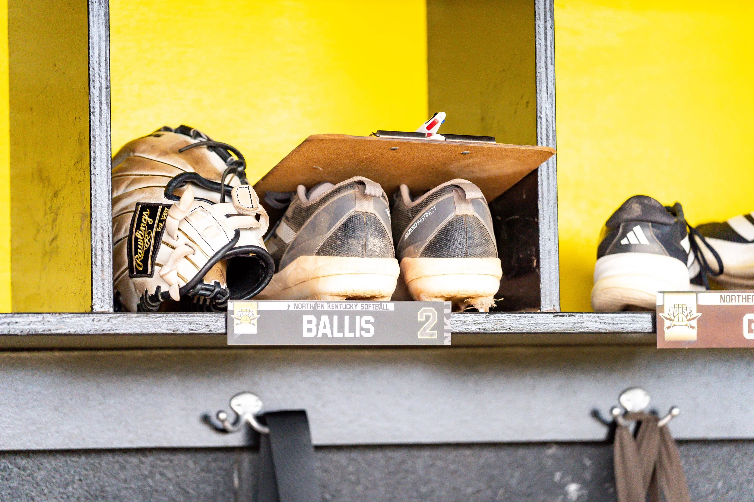 A baseball glove, shoes, and a clipboard on a shelf labeled 'BALLS' in a locker room with a yellow wall.
