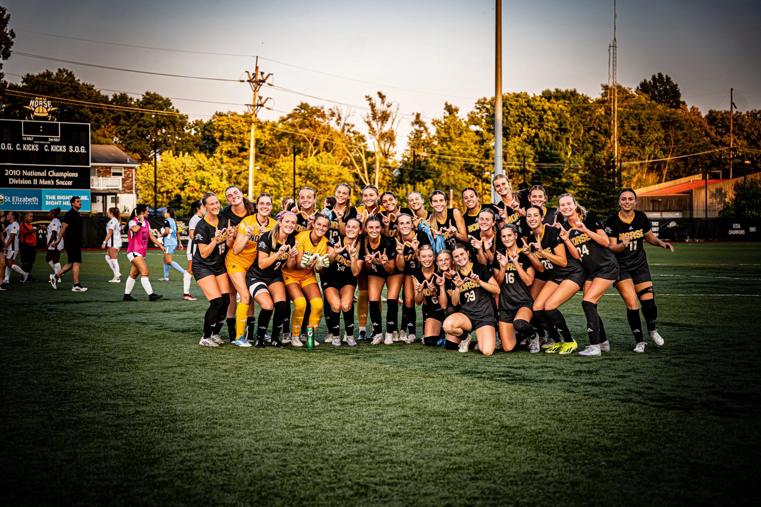 A group of female soccer players in black and yellow uniforms posing on a soccer field during sunset, smiling and making victory signs.