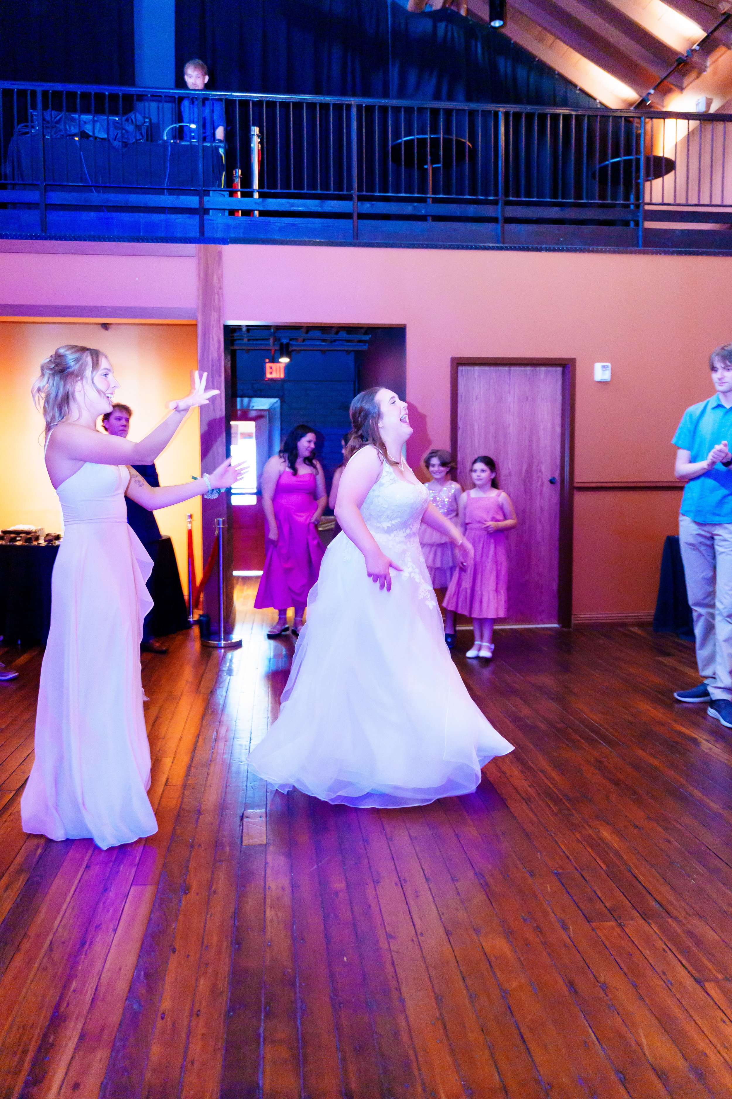 A woman in a white wedding dress is laughing and dancing at a wedding reception. She is surrounded by other women in pink dresses and a man in a blue shirt. The event is indoors with wooden flooring and warm orange walls.