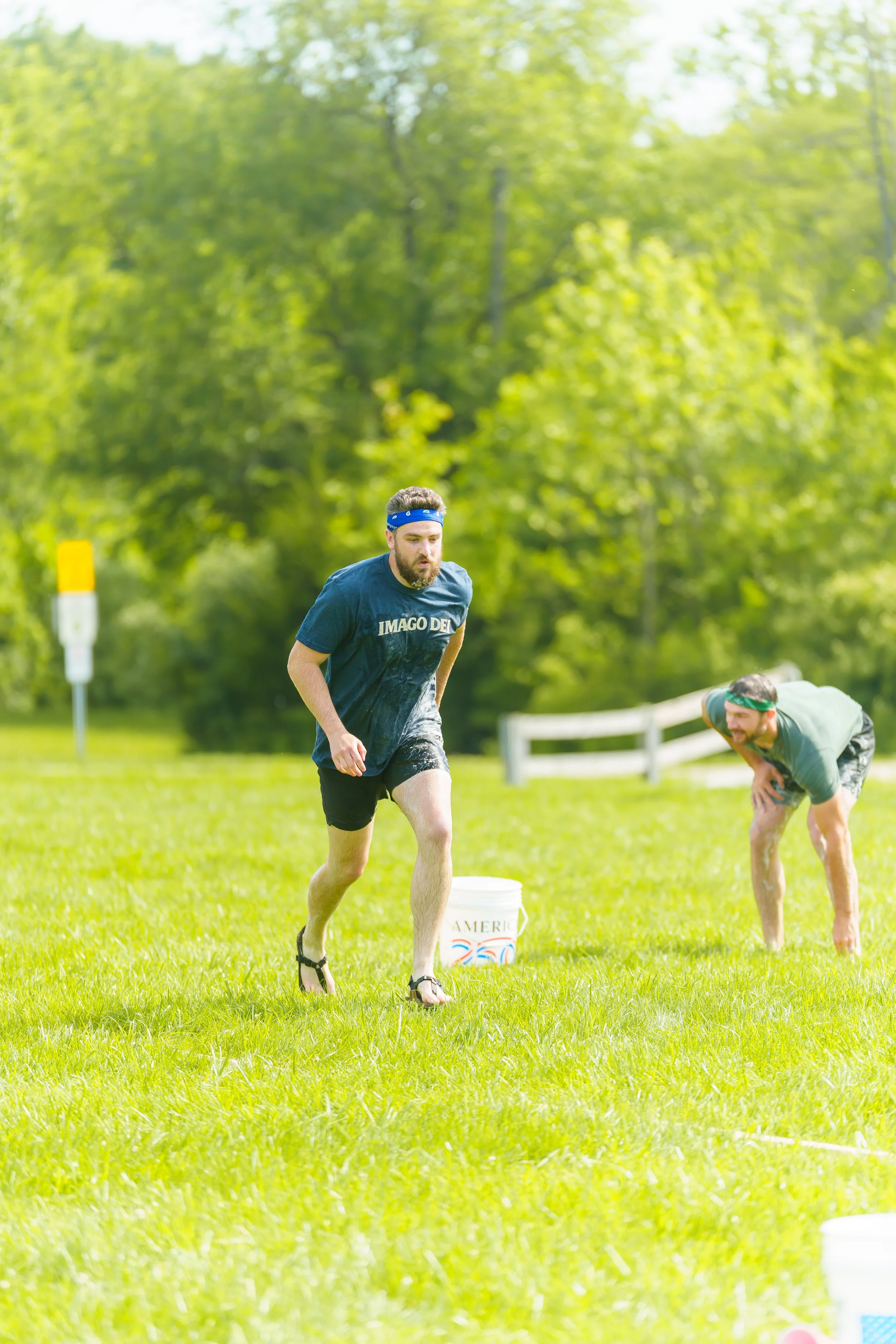 A man wearing a blue headband and a navy T-shirt running on a grassy field during daytime, with another person in the background also participating in an outdoor activity.