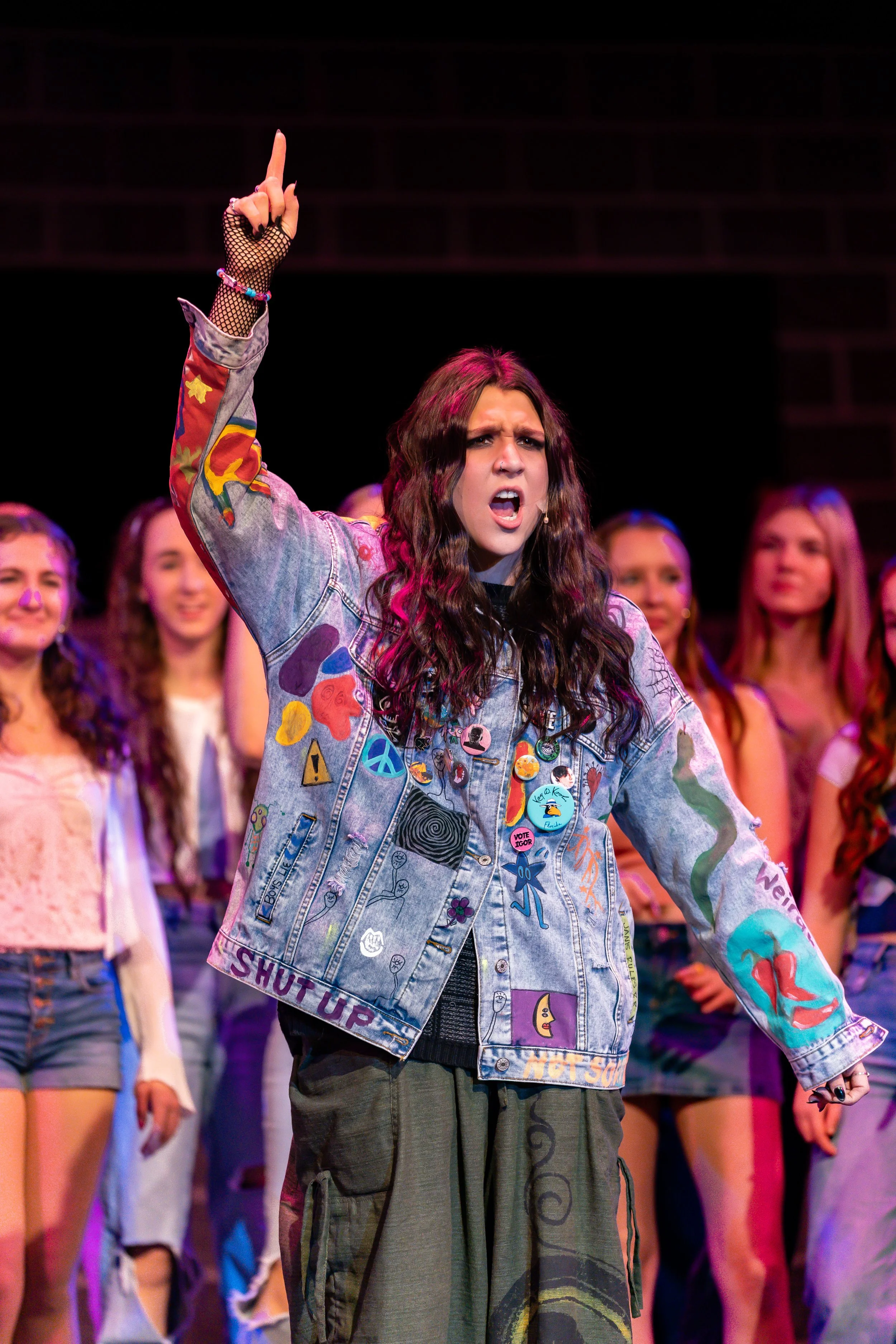 A young woman with long, wavy dark hair with pink highlights, wearing a decorated denim jacket and cargo pants, standing on stage with her middle finger raised and a frustrated facial expression. She is surrounded by other young women.