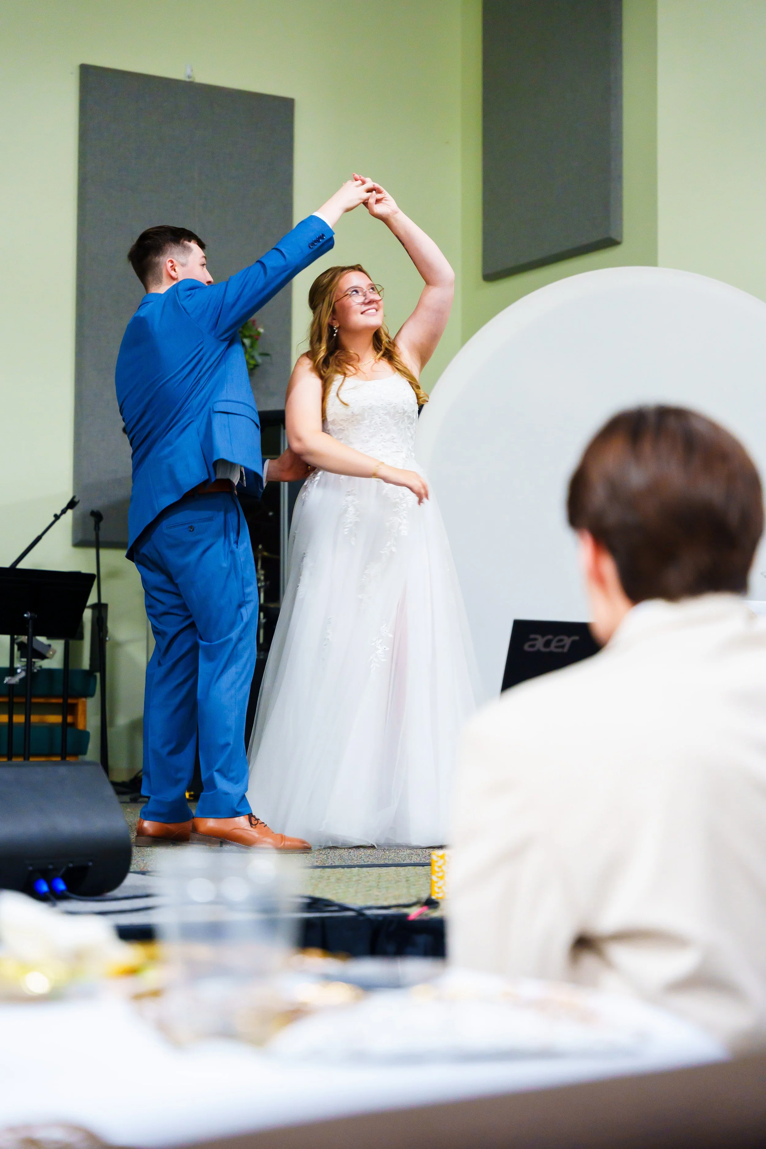 A couple dancing at a wedding ceremony on a stage, with guests seated in front. The groom is wearing a bright blue suit and brown shoes, and the bride is wearing a white wedding dress and glasses. The background includes a green wall and sound-absorb