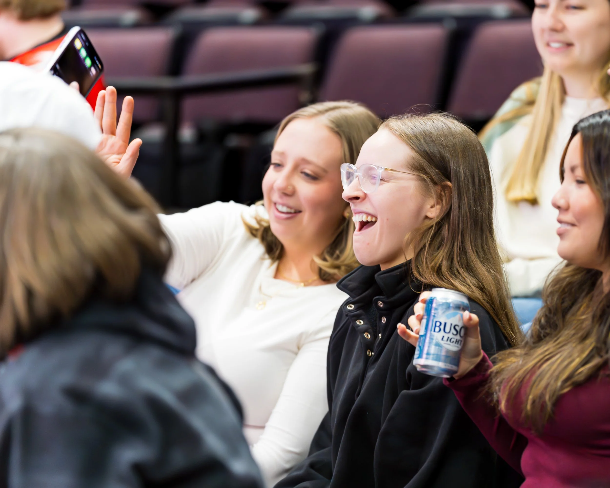 A group of young women sitting together, smiling and taking a selfie at what appears to be an indoor event or gathering, with empty chairs in the background.