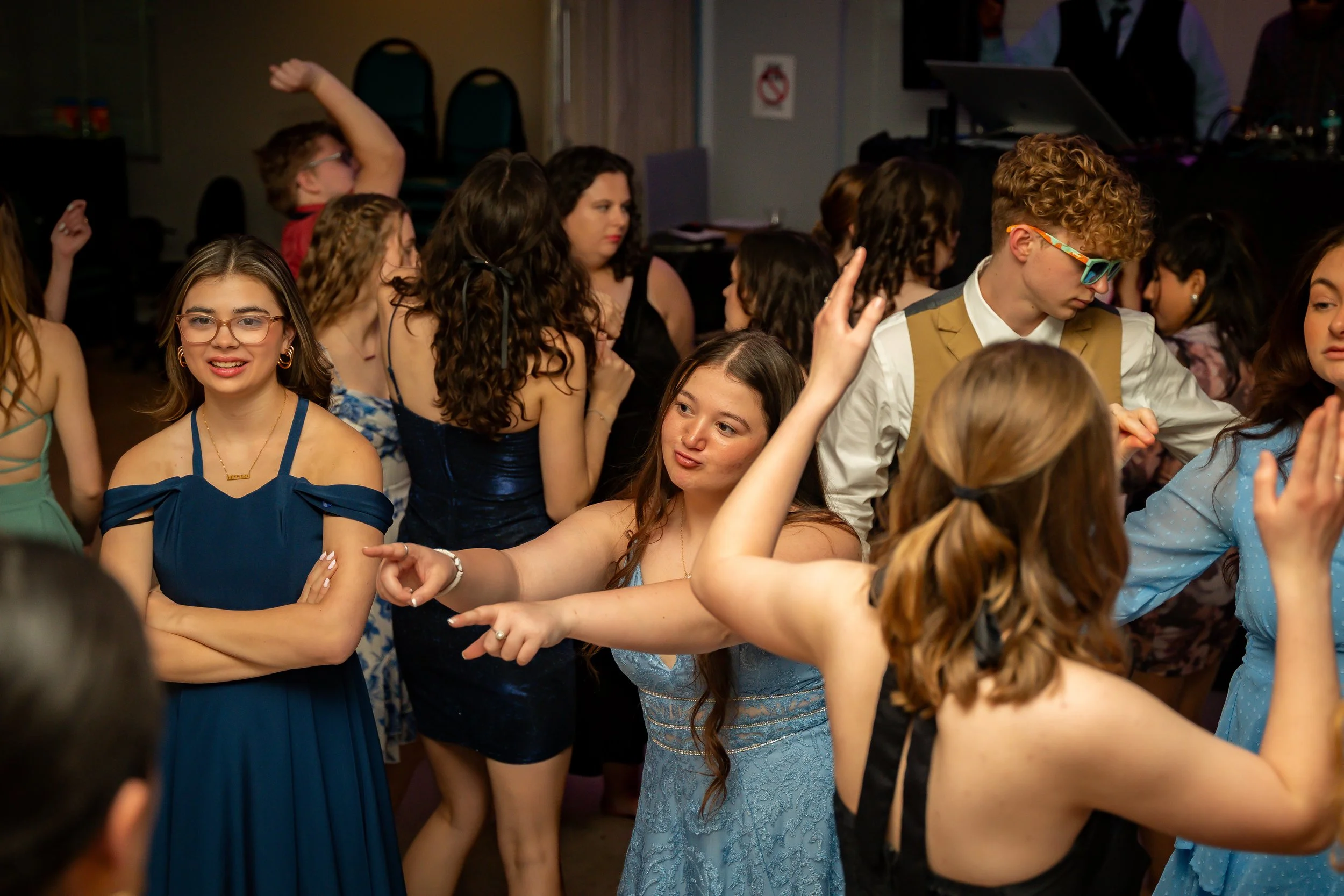 A group of young women and a young man dancing and enjoying a social event in a dimly lit room, some raising their hands and smiling.