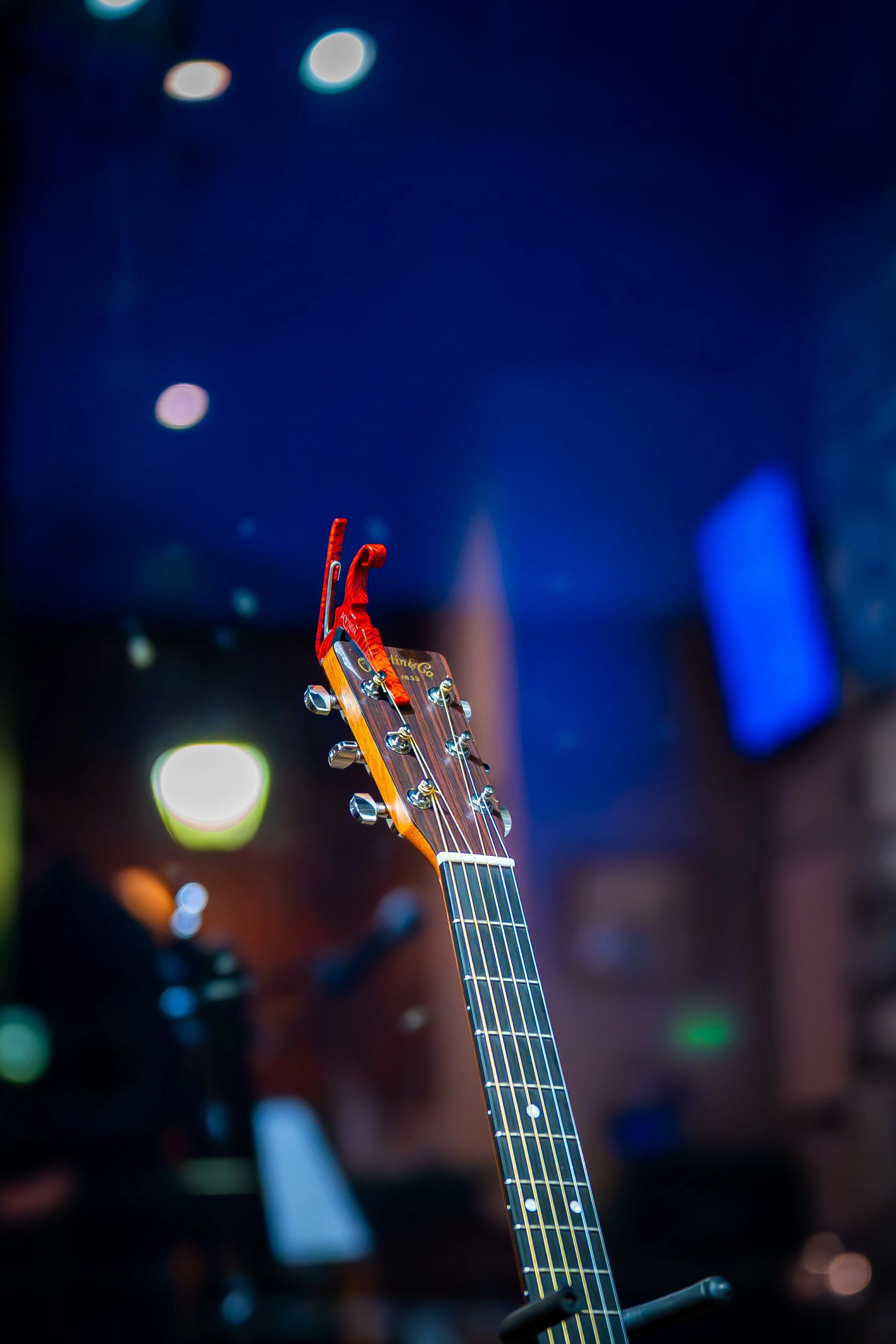 Close-up of an acoustic guitar headstock with tuning pegs, set against a dark background with colorful bokeh lights.