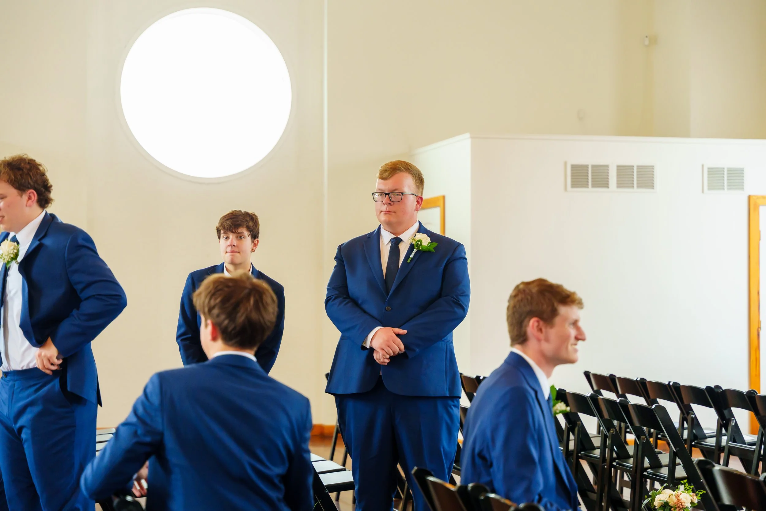 A group of young men dressed in blue suits with white shirts and dark ties, standing in a room during a formal event, possibly a wedding or ceremony.