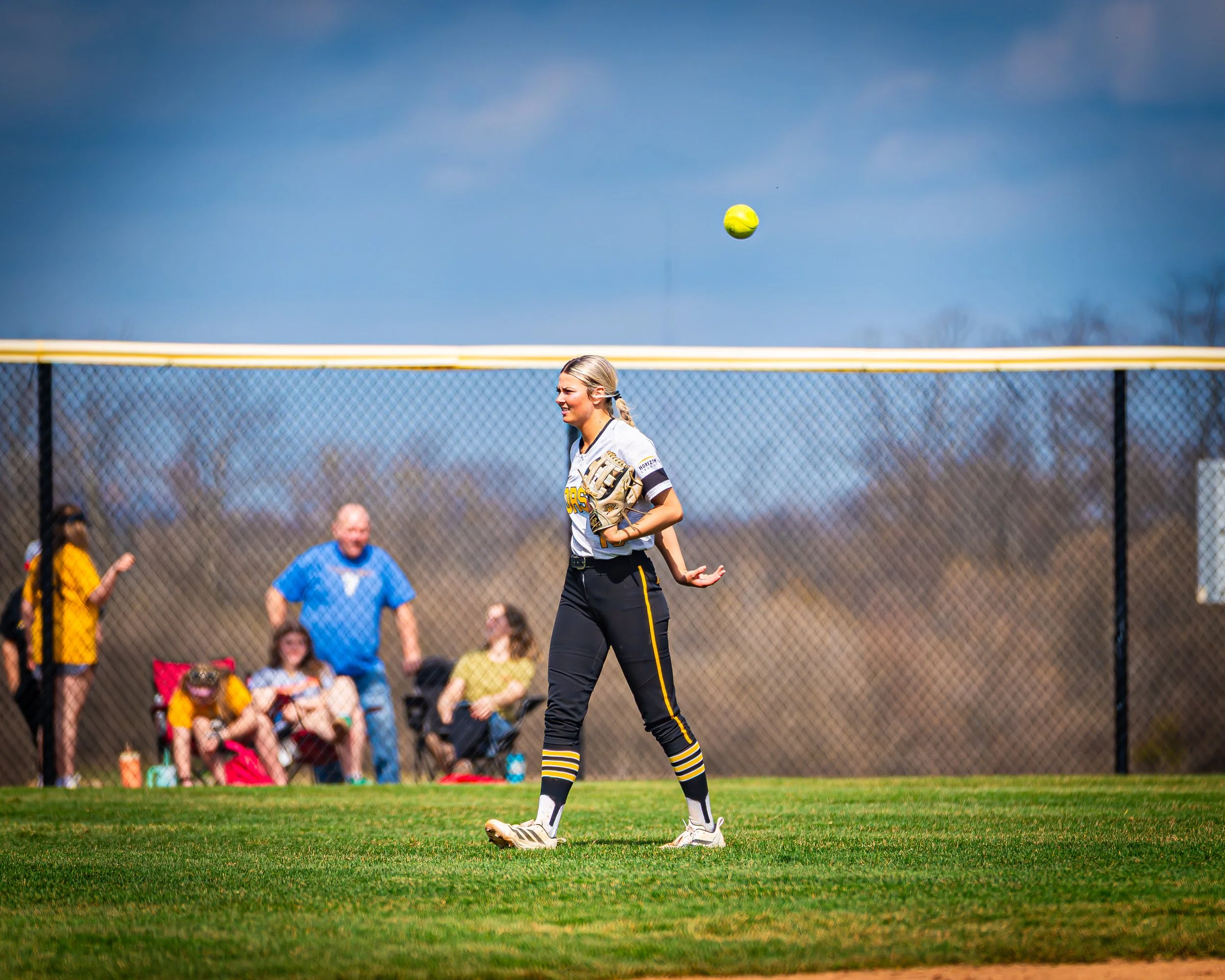 A female softball player in a gray and black uniform with yellow accents, standing on a grassy field, preparing to catch a yellow softball in midair. Spectators are sitting behind a chain-link fence, and a blue sky is visible above.