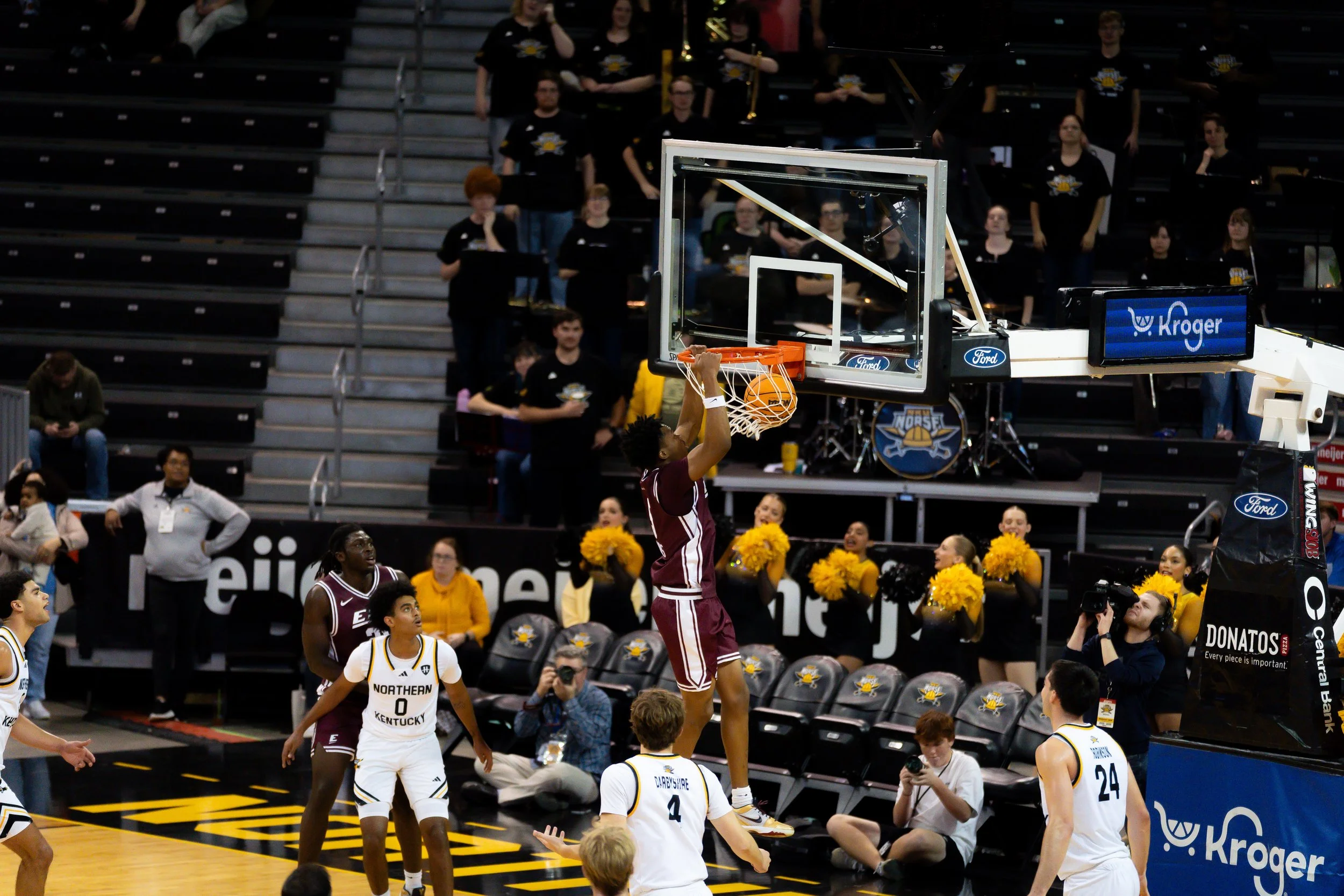 A college basketball game with players in white and maroon jerseys. A player in maroon jumps to make a dunk, while players in white watch or prepare to coordinate. The court has black and yellow accents, and cheerleaders in yellow pom-poms are seated