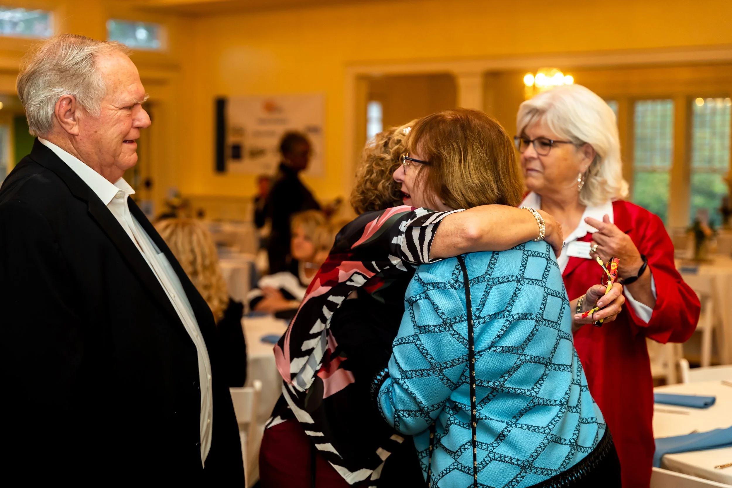 A group of people at a social gathering, with one man in a black suit on the left and two women on the right embracing each other, one in a colorful dress and the other in a red jacket.