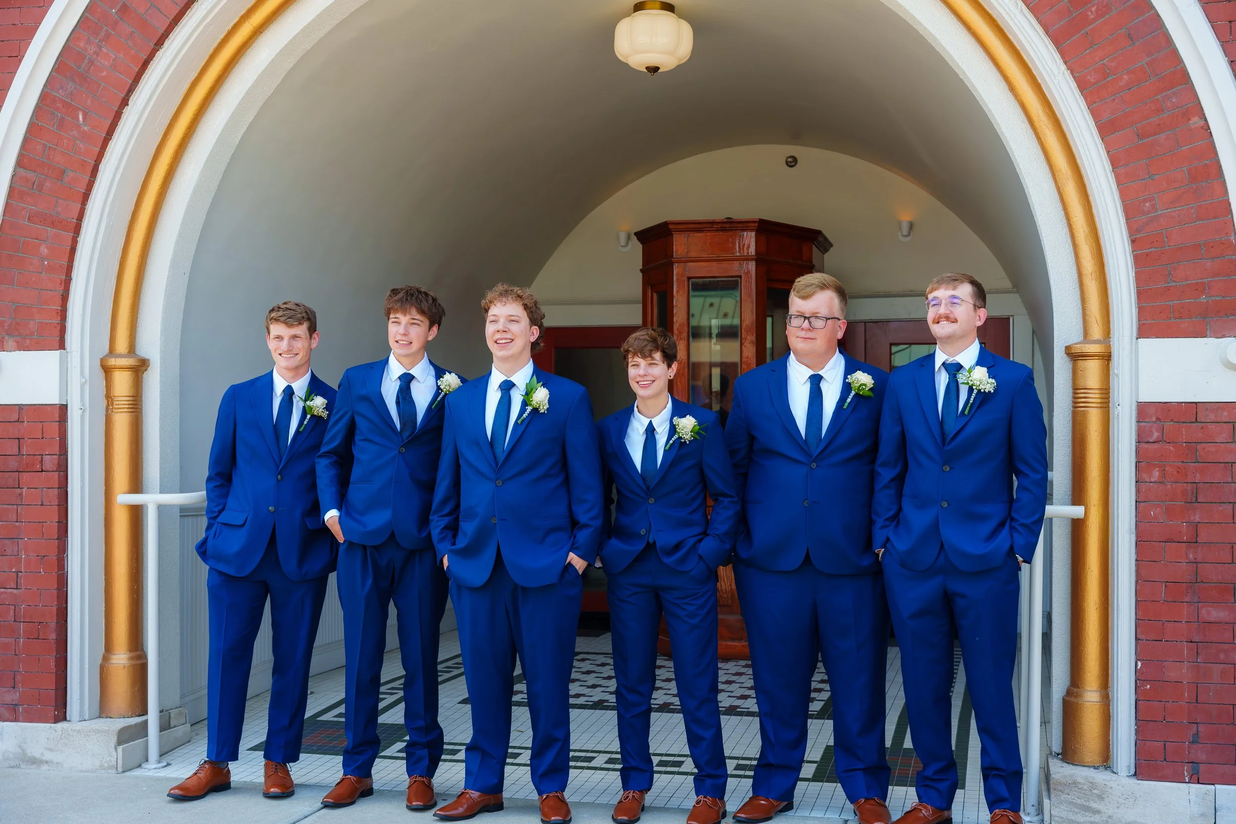 Group of six young men in matching blue suits with white shirts and blue ties standing under an arched doorway with brick and white trim, smiling, some with hands in pockets, celebrating a special occasion.