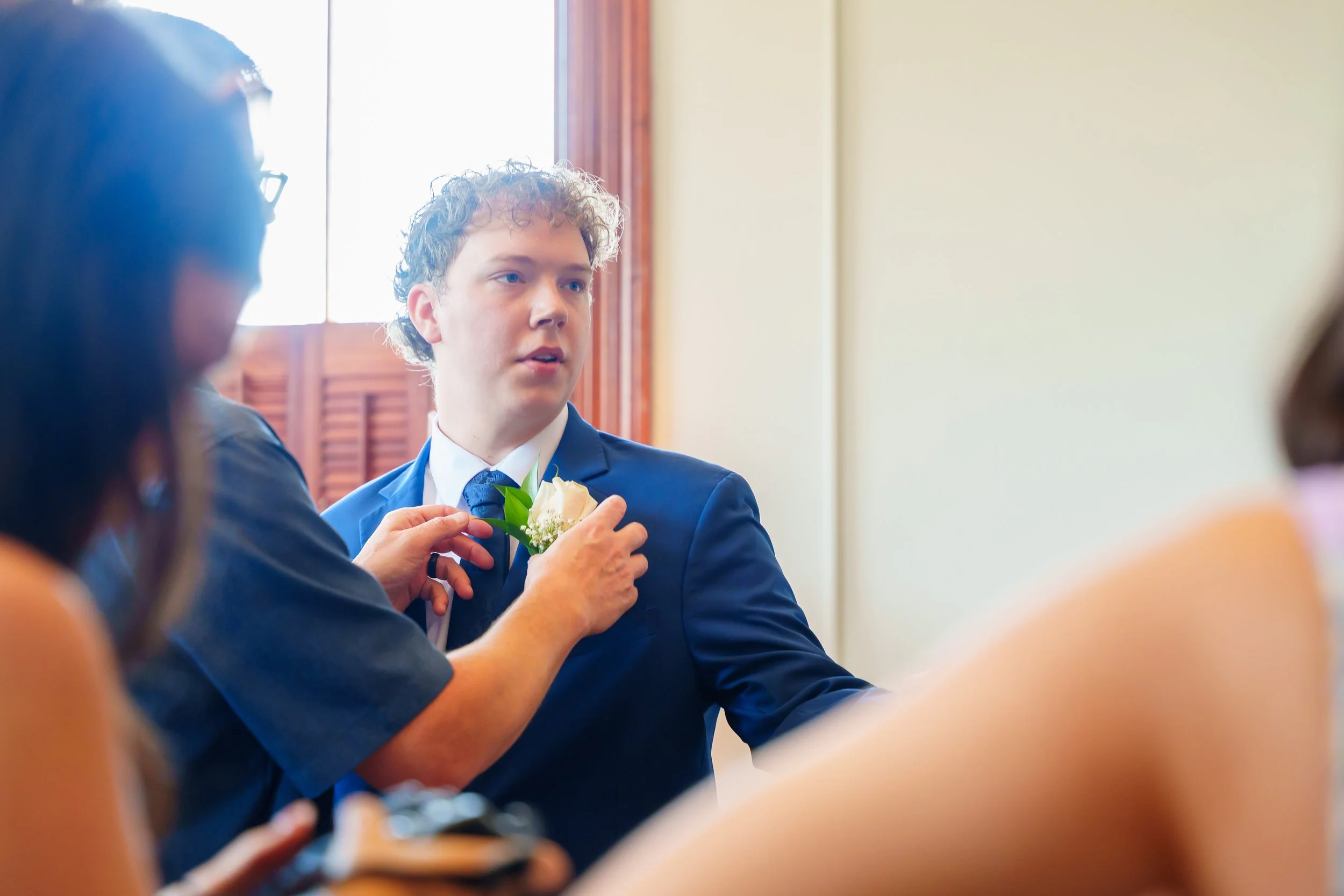 A young man in a suit and tie with curly hair receiving a white flower corsage during a formal event, with other people around him in a room with wooden window shutters.