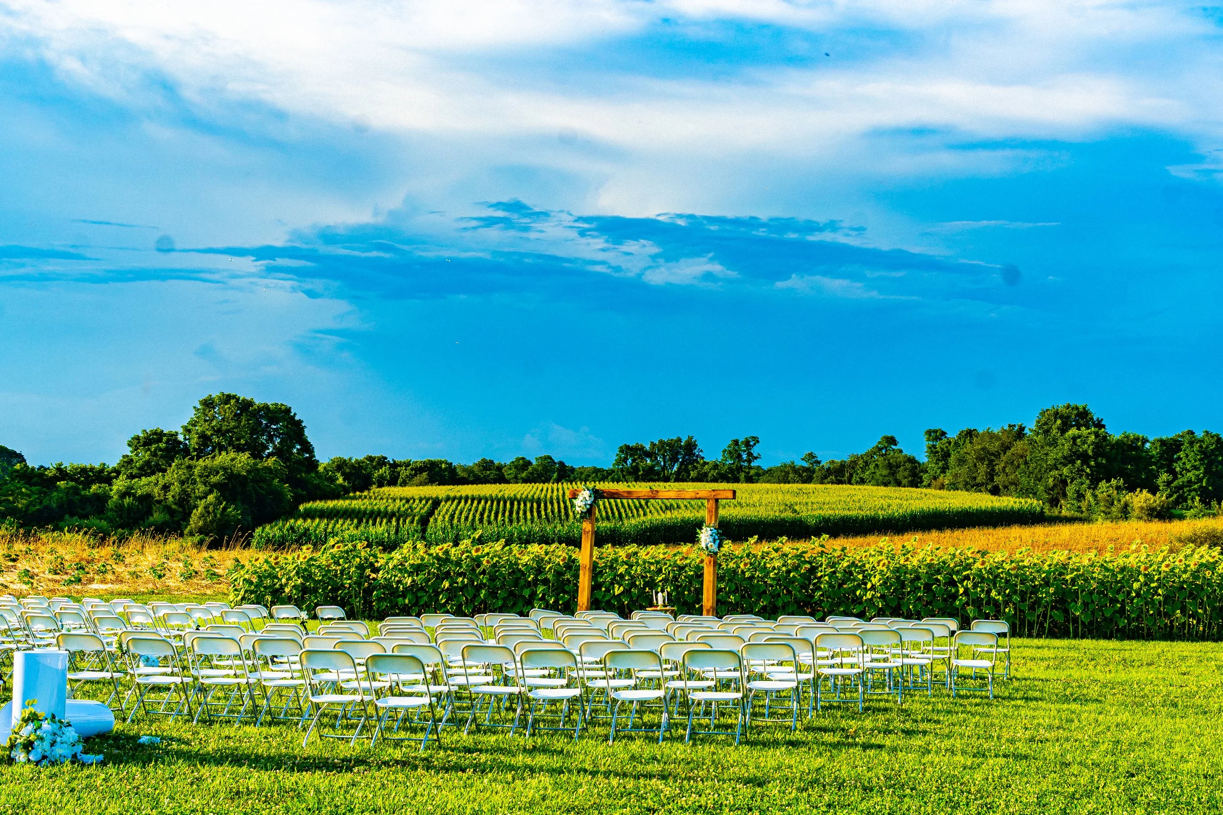 Outdoor wedding ceremony setup with rows of chairs on a grassy field, decorated with flowers, against a backdrop of a large green farm field, trees, and a partly cloudy blue sky.