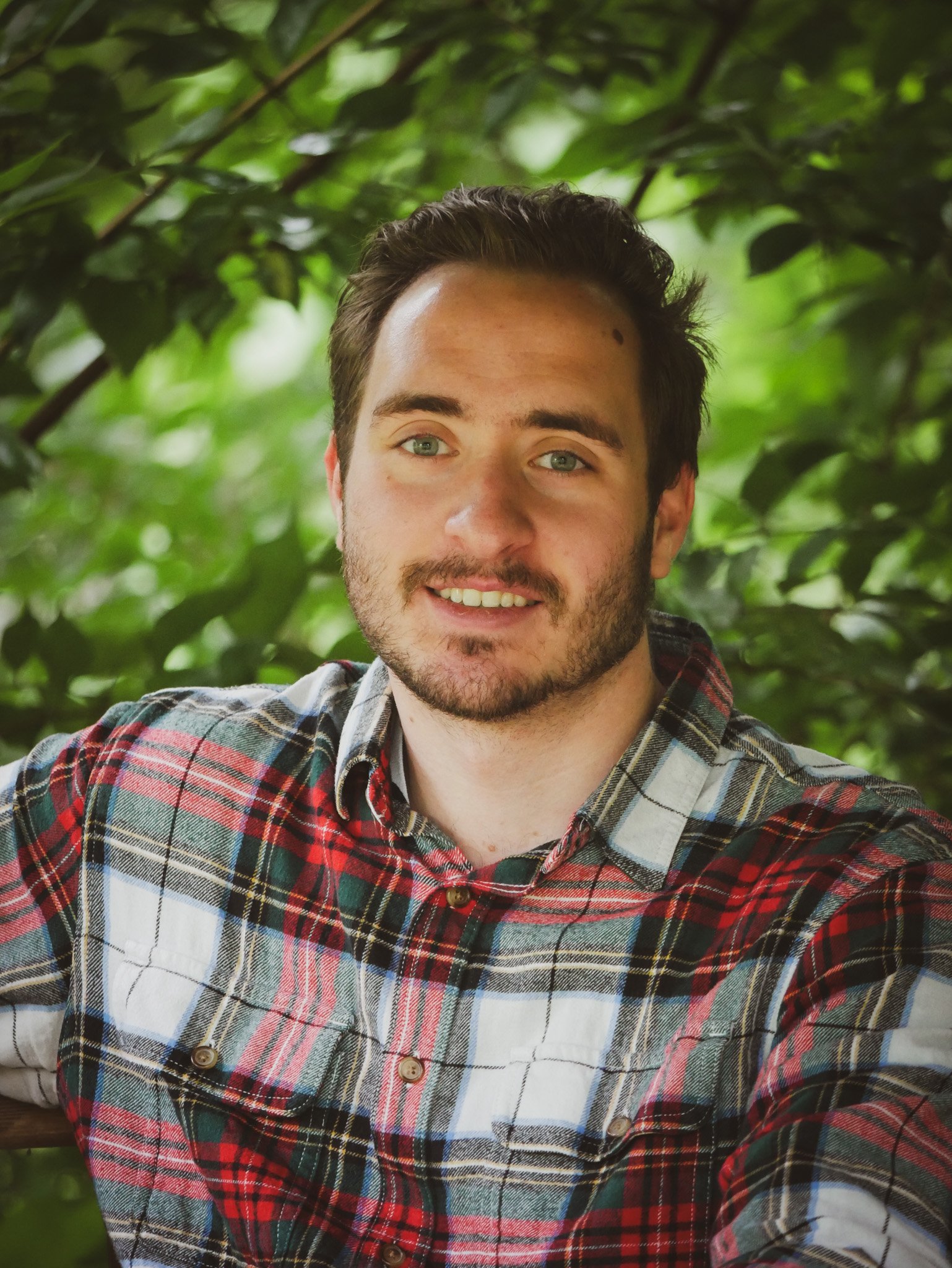 A young man with short dark hair, light beard, and light blue eyes smiling, wearing a plaid shirt with red, green, white, and yellow patterns, outdoors surrounded by green leaves.