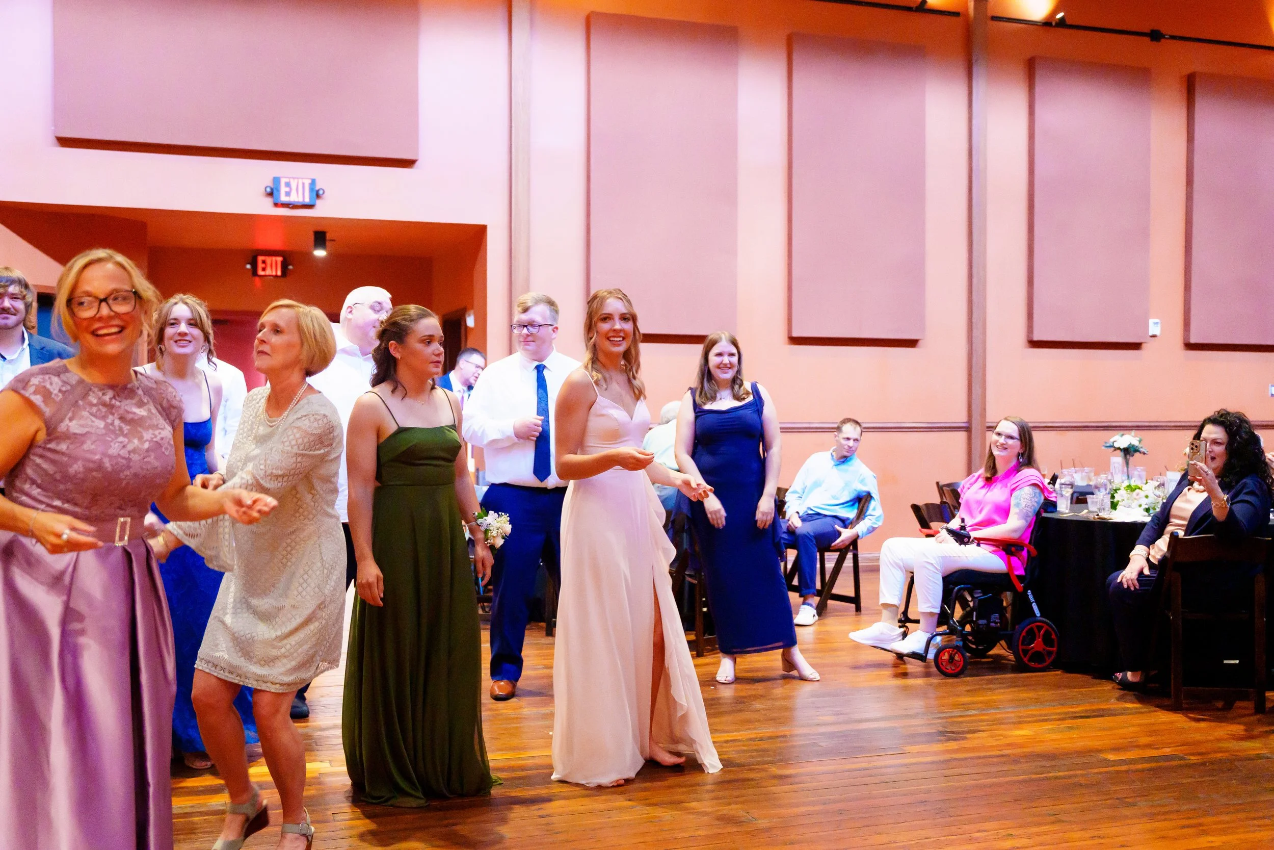 Group of women and men dressed in formal attire at a celebration or wedding reception in a hall with peach-colored walls and wooden floor.