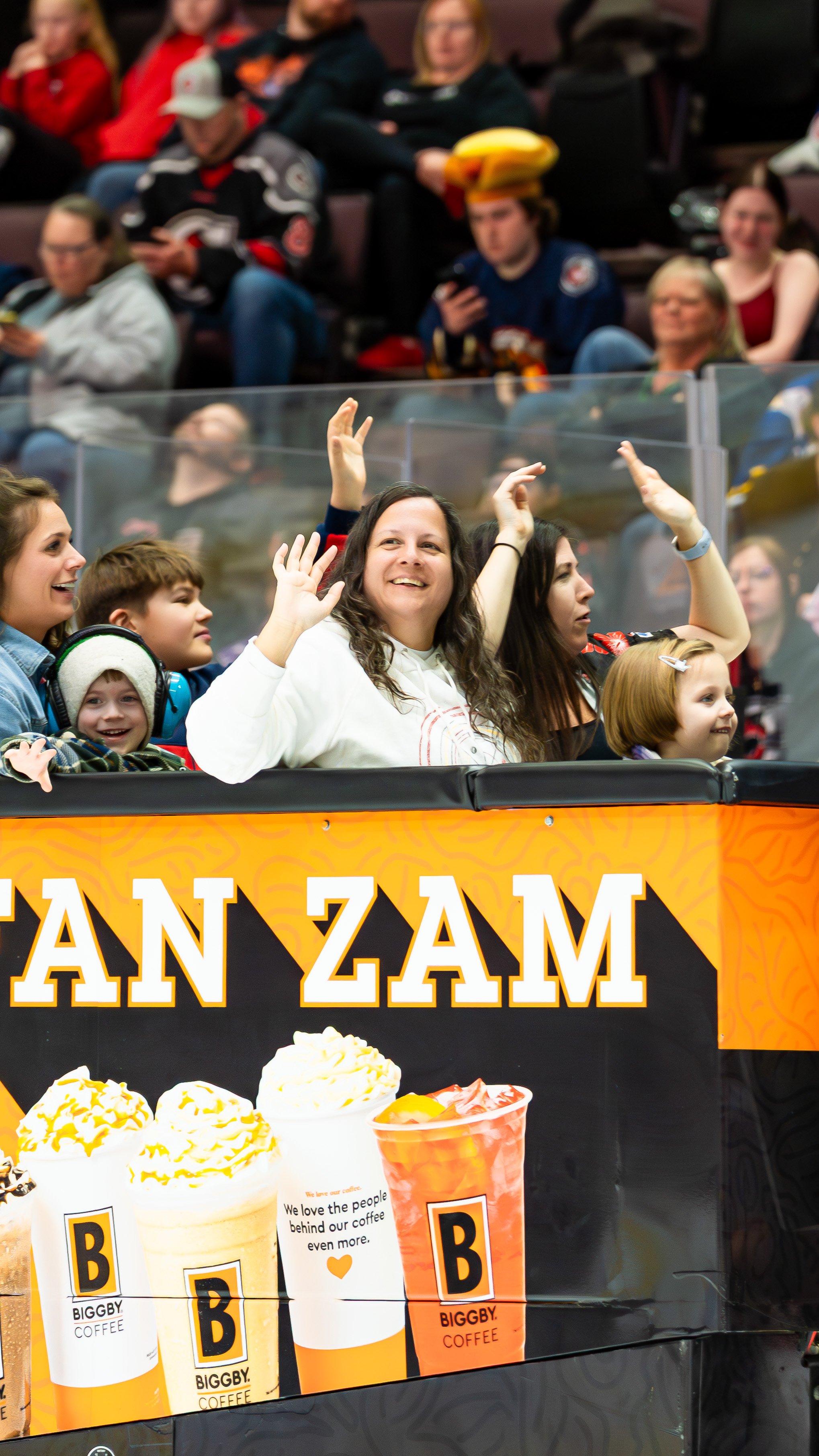 People sitting and watching an event at an ice hockey game, including children and adults, with a woman in the center waving and smiling.