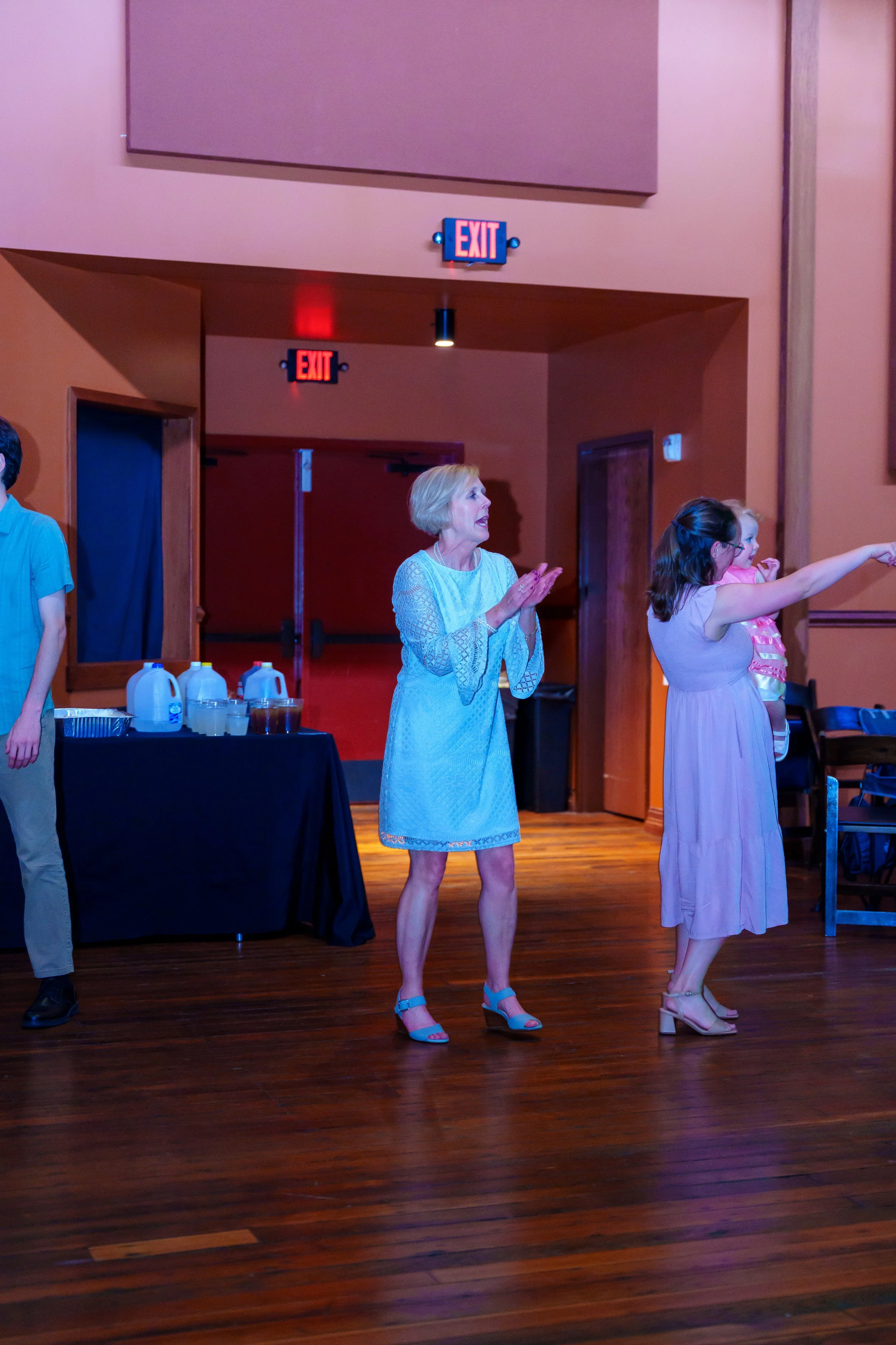 A woman and a girl standing in a room with wooden floors and walls, with two illuminated exit signs overhead. The woman is wearing a light blue dress and teal high heels, and the girl is wearing a pink dress and beige high heels. The girl is pointing