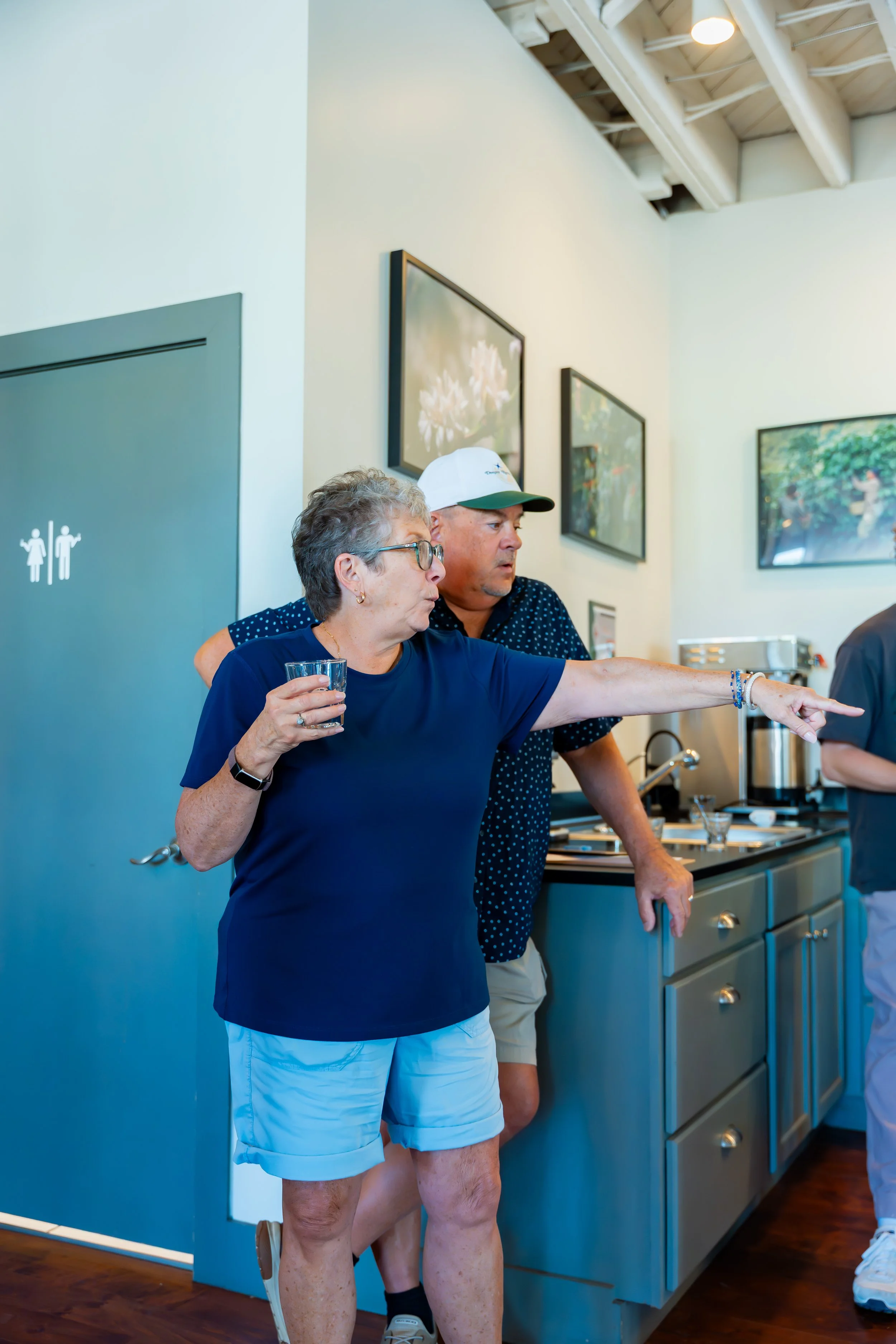 Two women standing together in a room, one pointing towards something outside the frame, with a kitchen counter and framed artwork on the wall behind them.