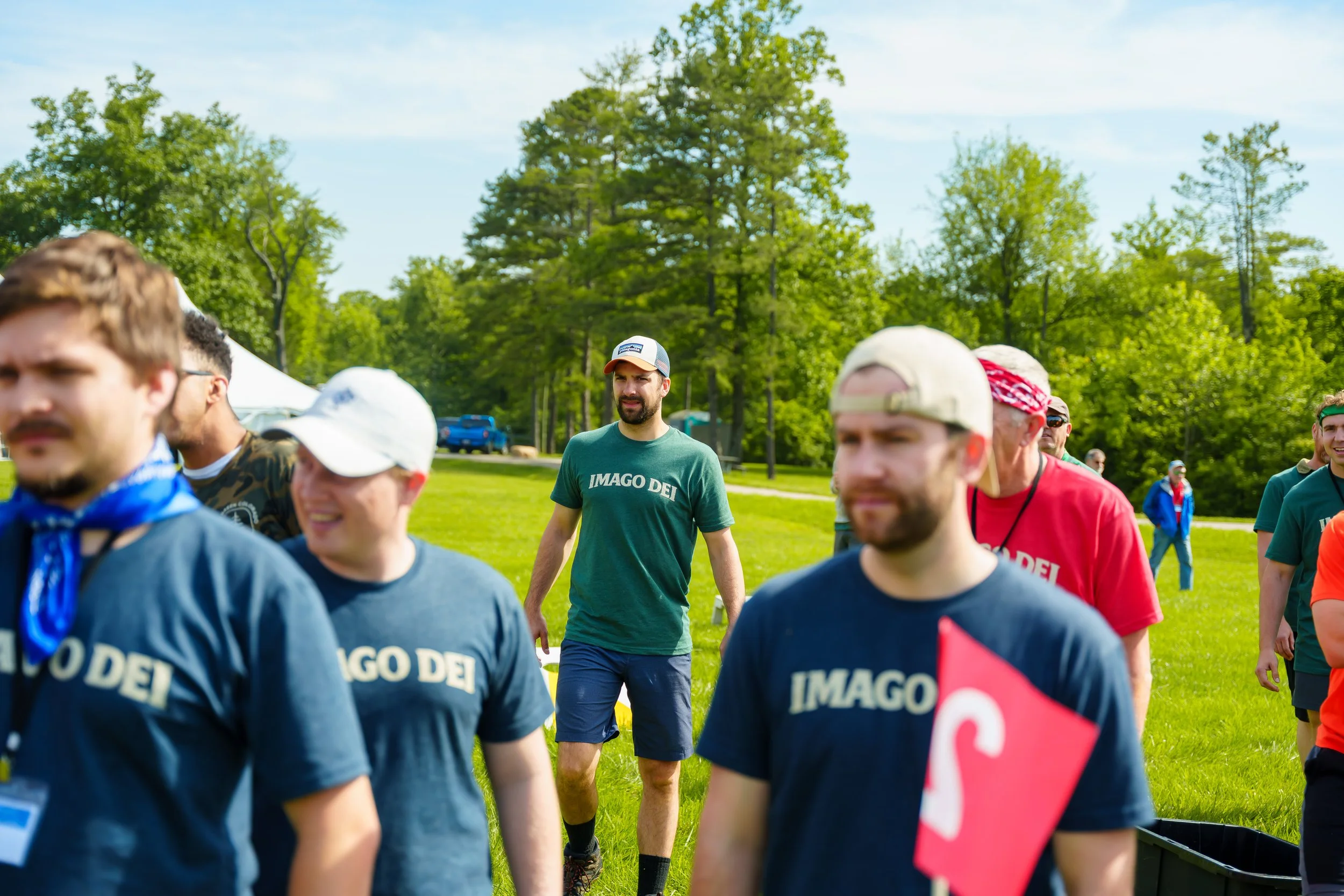 Group of young people walking outdoors in a park on a sunny day, some wearing caps and t-shirts that say 'IMAGO DEI', surrounded by green trees and grass.