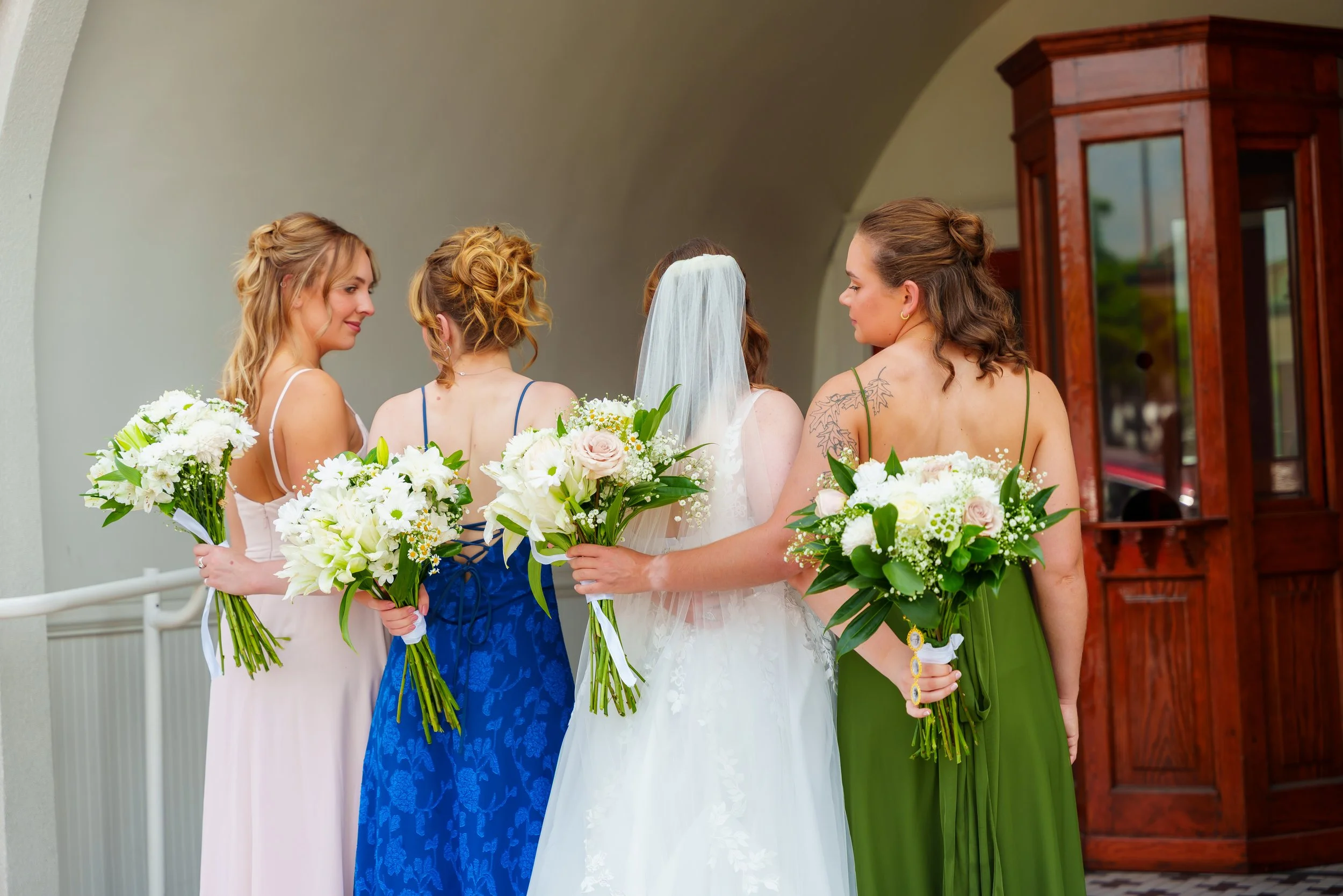 Group of women with bouquets standing together at a wedding, one woman in a bridal gown and veil, others in colorful dresses, outside near a building with a wooden door.
