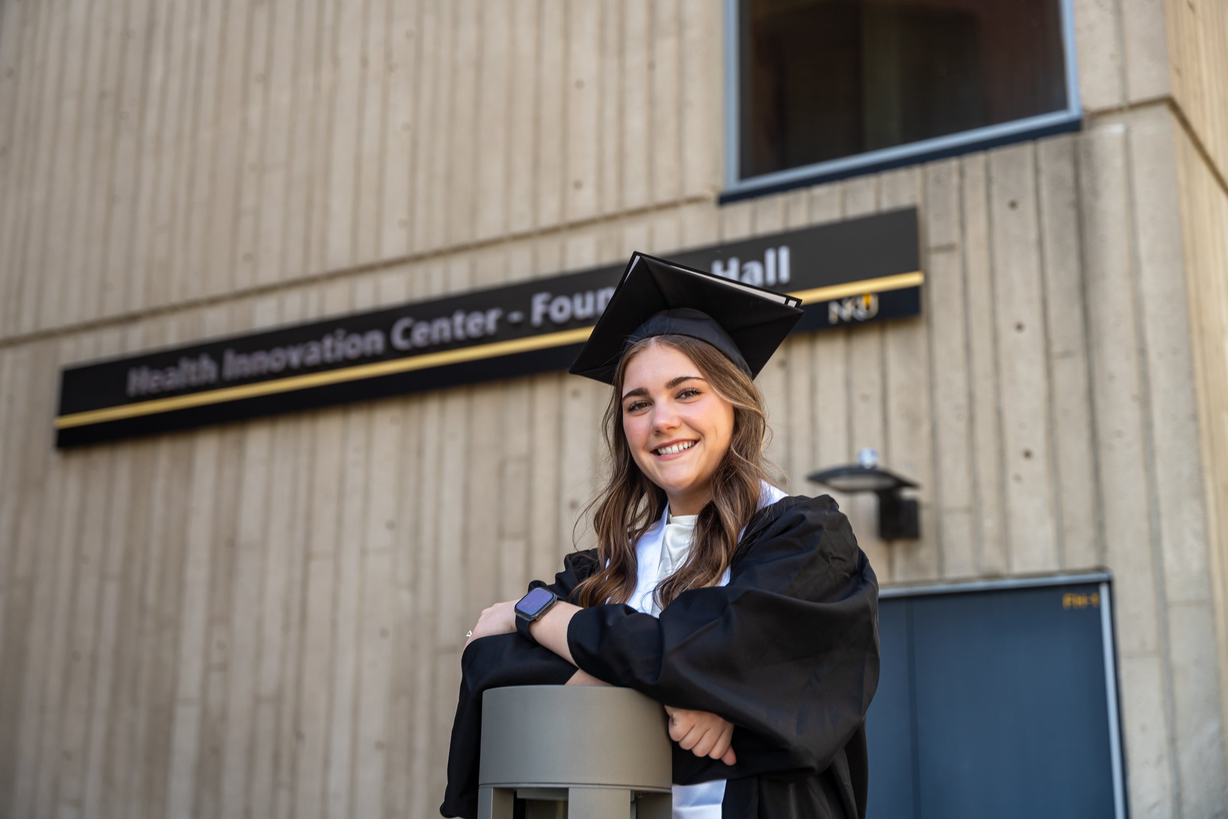 A young woman in graduation gown and cap smiling while crossing her arms, standing outdoors in front of a building with a sign that reads 'Health Innovation Center.'