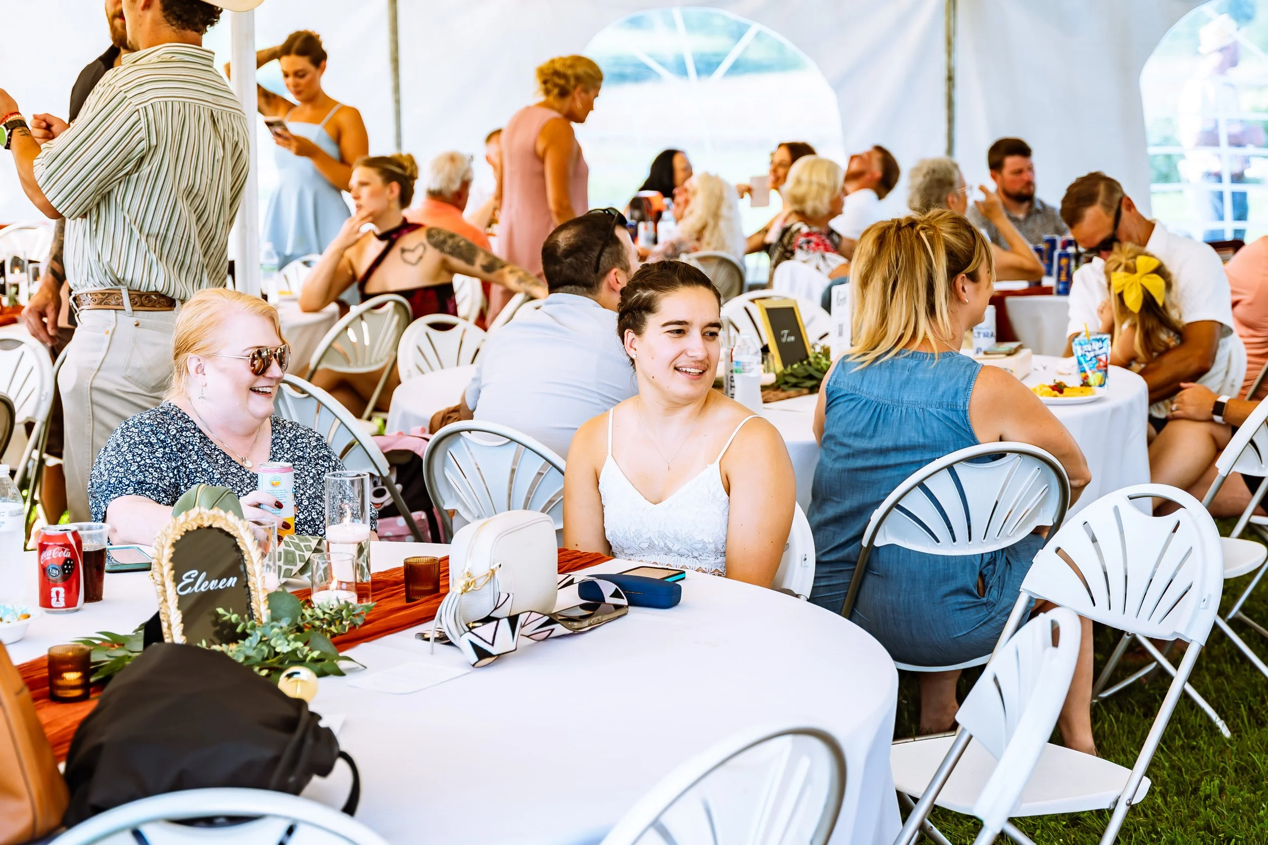People gathered at a daytime outdoor celebration dining under a white tent, with tables decorated with greenery, flowers, and place cards.