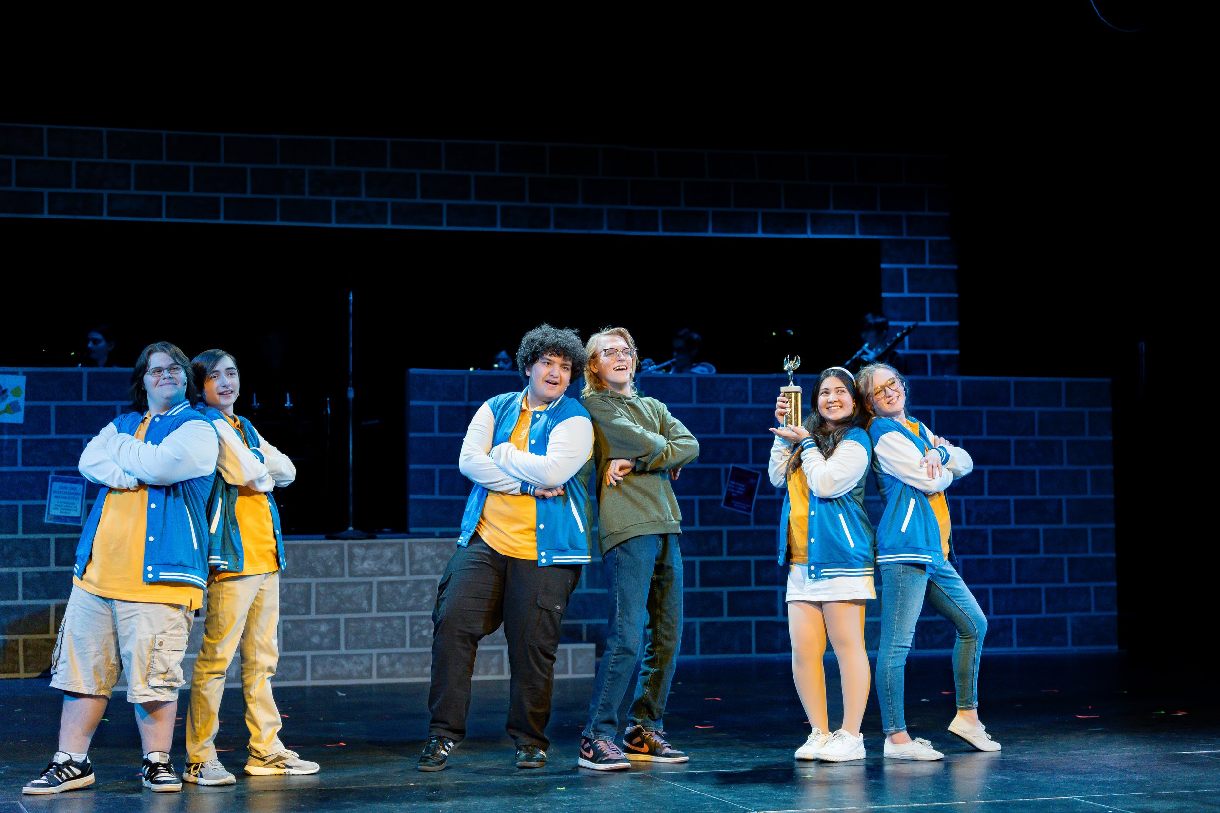 Group of six diverse teenagers on stage, five wearing yellow and blue jackets and one with glasses holding a trophy, with a dark background and blue brick wall.