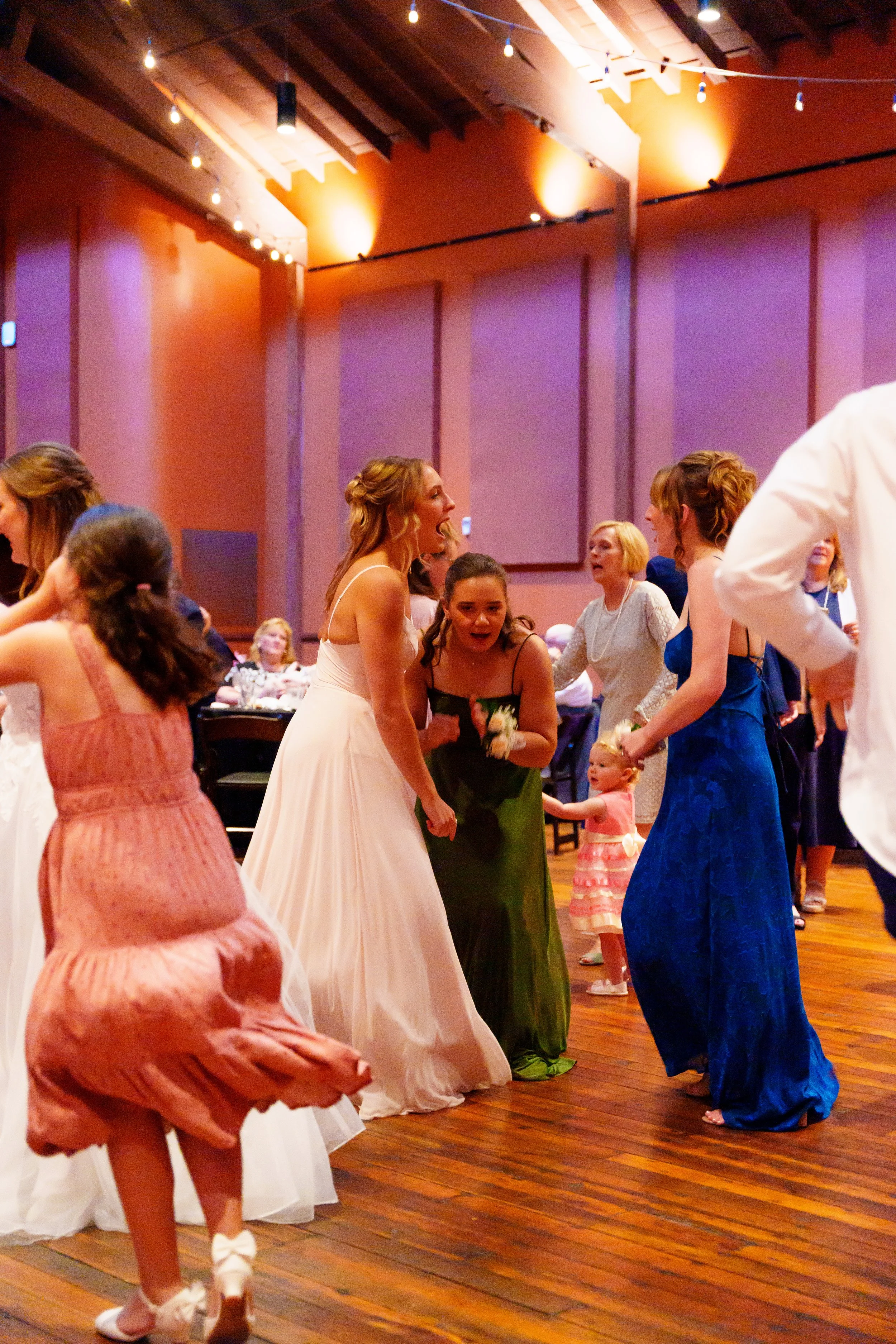 A group of women and children dancing and socializing at a wedding reception in a decorated hall with purple walls and wooden floors. The women are dressed in colorful gowns, and some children are in dresses. The scene is lively and joyous.