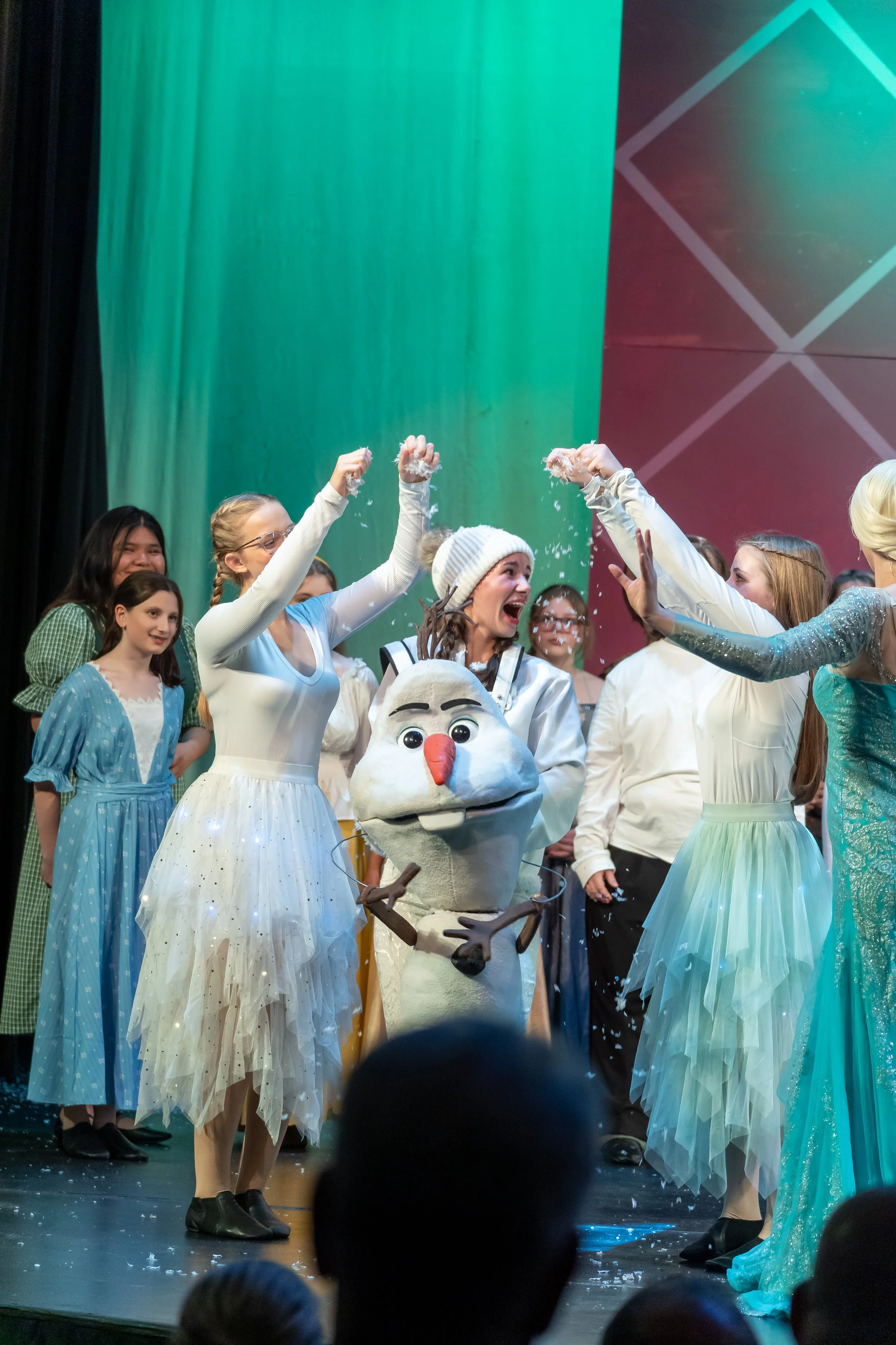 A group of people, mostly wearing colorful dresses, celebrating on stage with a costumed Olaf character from Frozen, while confetti falls around them.