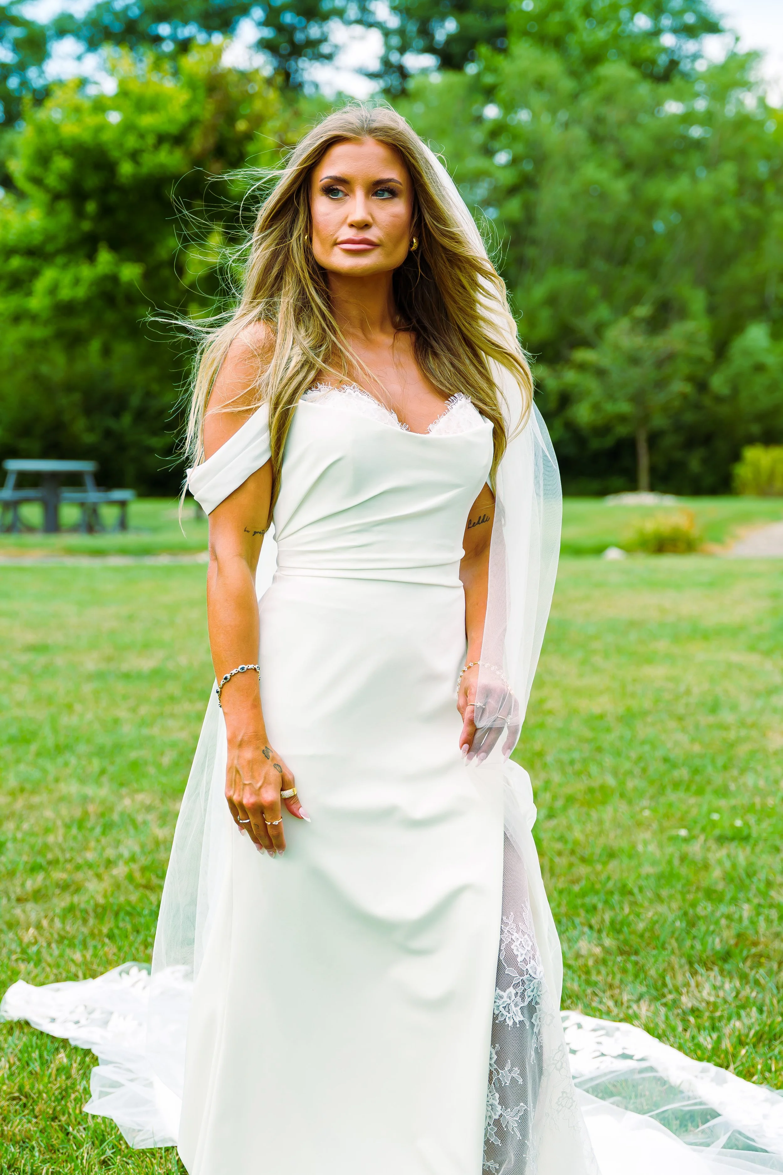 A woman in a white wedding dress standing outdoors in a grassy area with trees in the background.