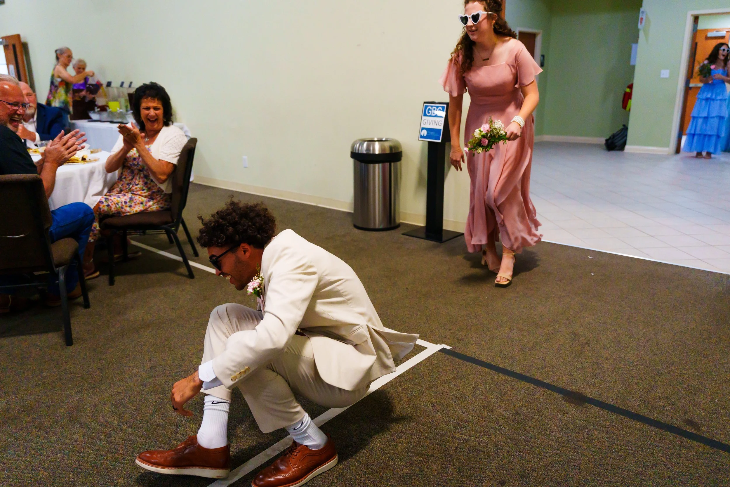 A man in a white suit and brown shoes is bending down and pretending to tie his shoelace while on a dance floor. A woman in a pink dress and heart-shaped sunglasses is walking past him with a small bouquet of flowers. In the background, seated guests